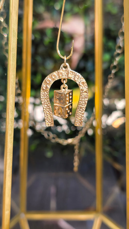 A gold-toned horseshoe-shaped pendant with a small rectangular charm featuring a raised '1', hanging in front of a blurred gold display case and green foliage background.