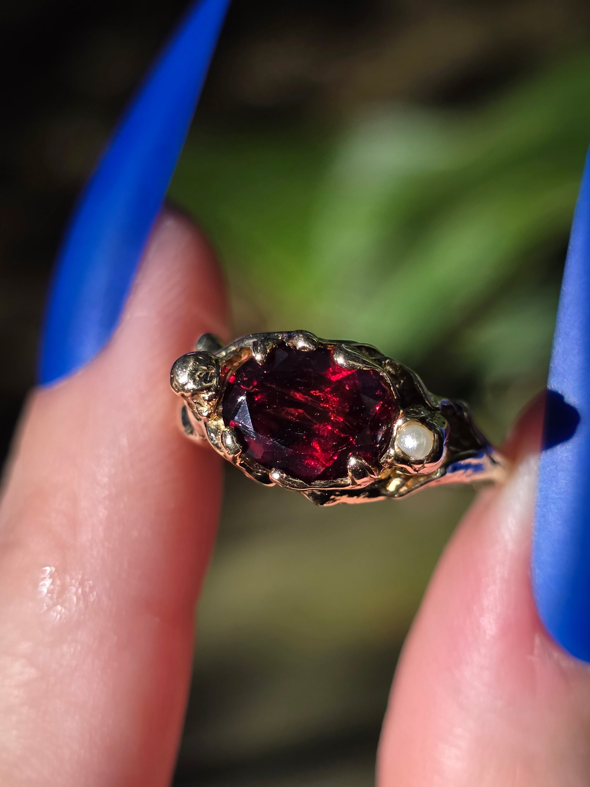 Close-up of a gold ring with a red gemstone held between fingers against a blurred green background