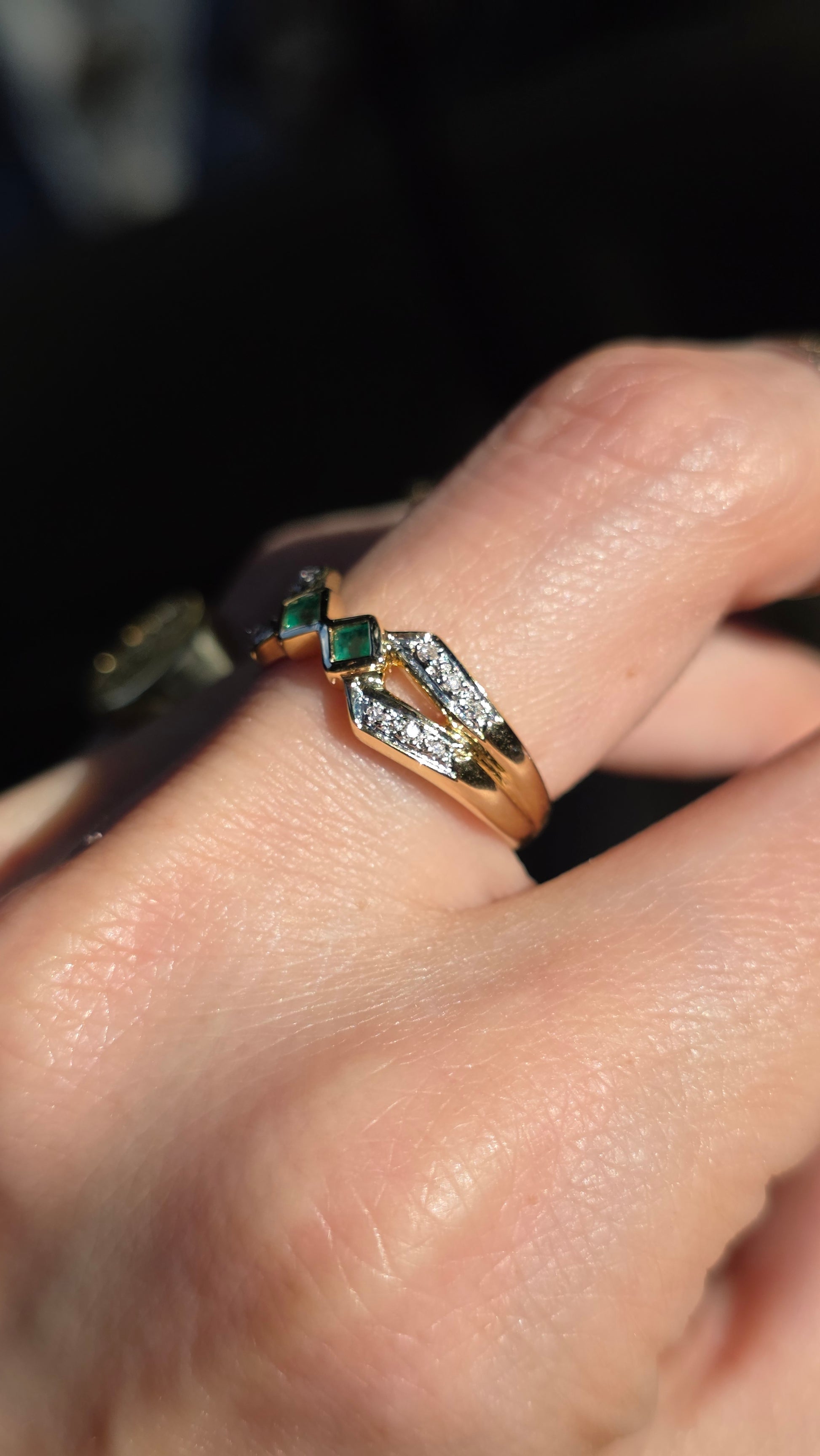 A close-up of a hand wearing a gold ring with a geometric design set with small clear stones and green square emerald-cut accents.