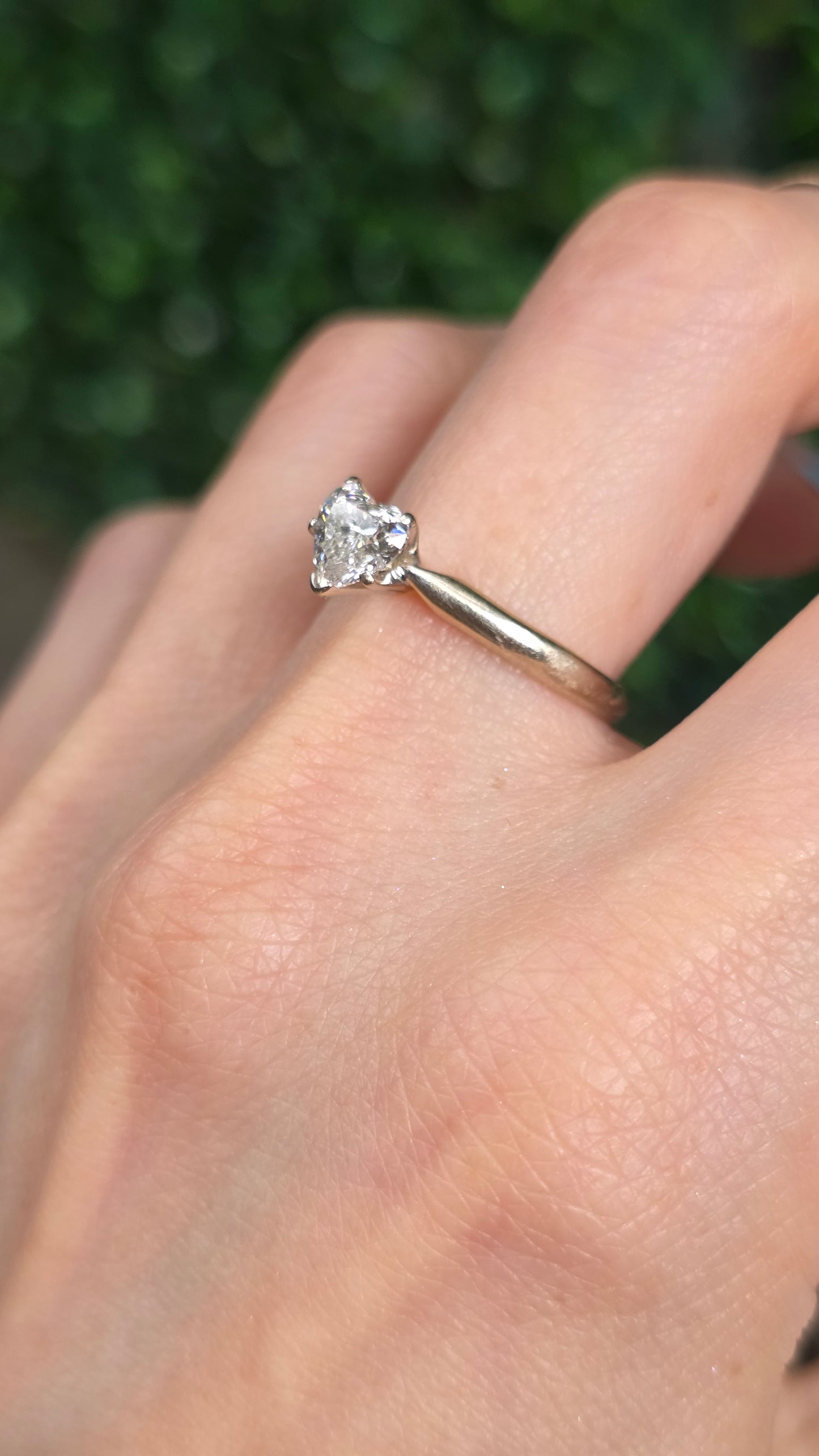 A close-up of a hand wearing a thin gold band ring with a small clear heart-shaped gemstone, set against a blurred green background.