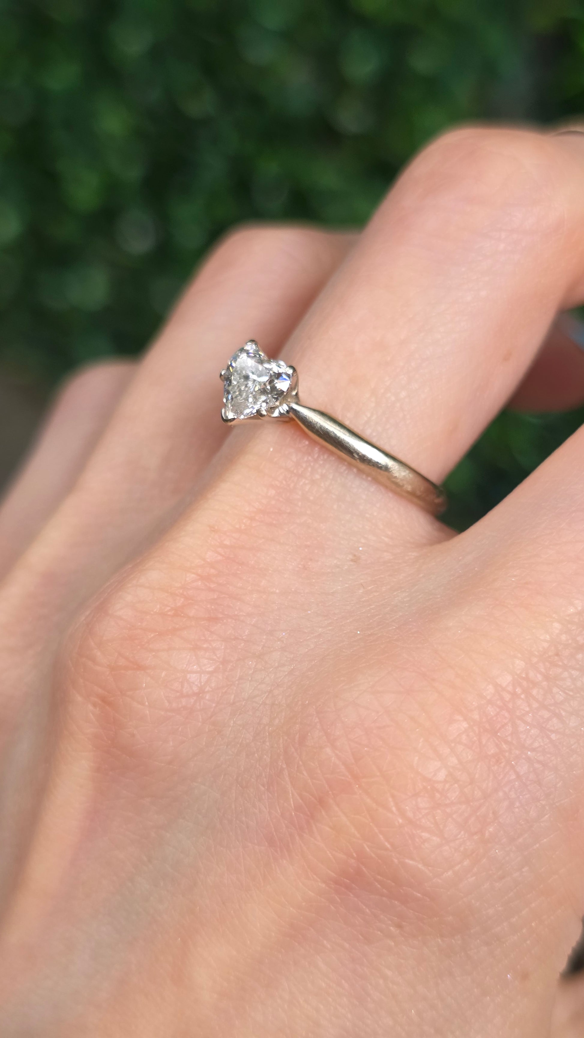 A close-up of a hand wearing a thin gold band ring with a small clear heart-shaped gemstone, set against a blurred green background.