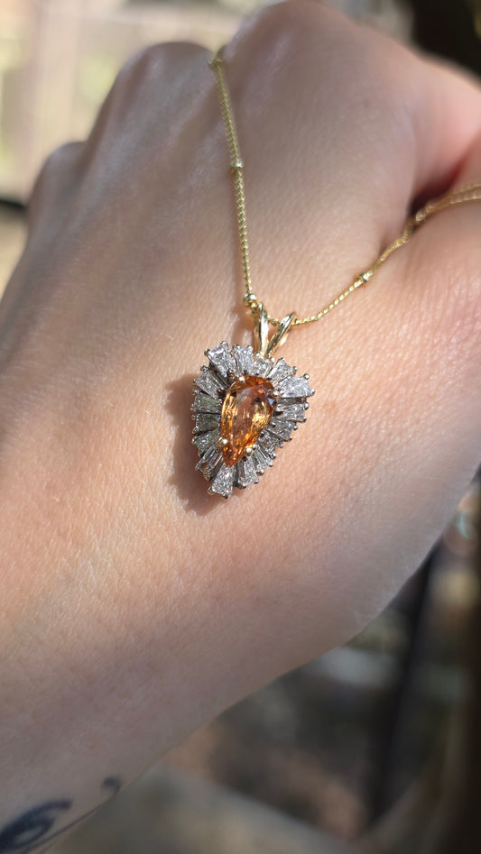 A close-up of a hand wearing a gold pendant necklace with a teardrop-shaped orange gemstone surrounded by small clear stones.