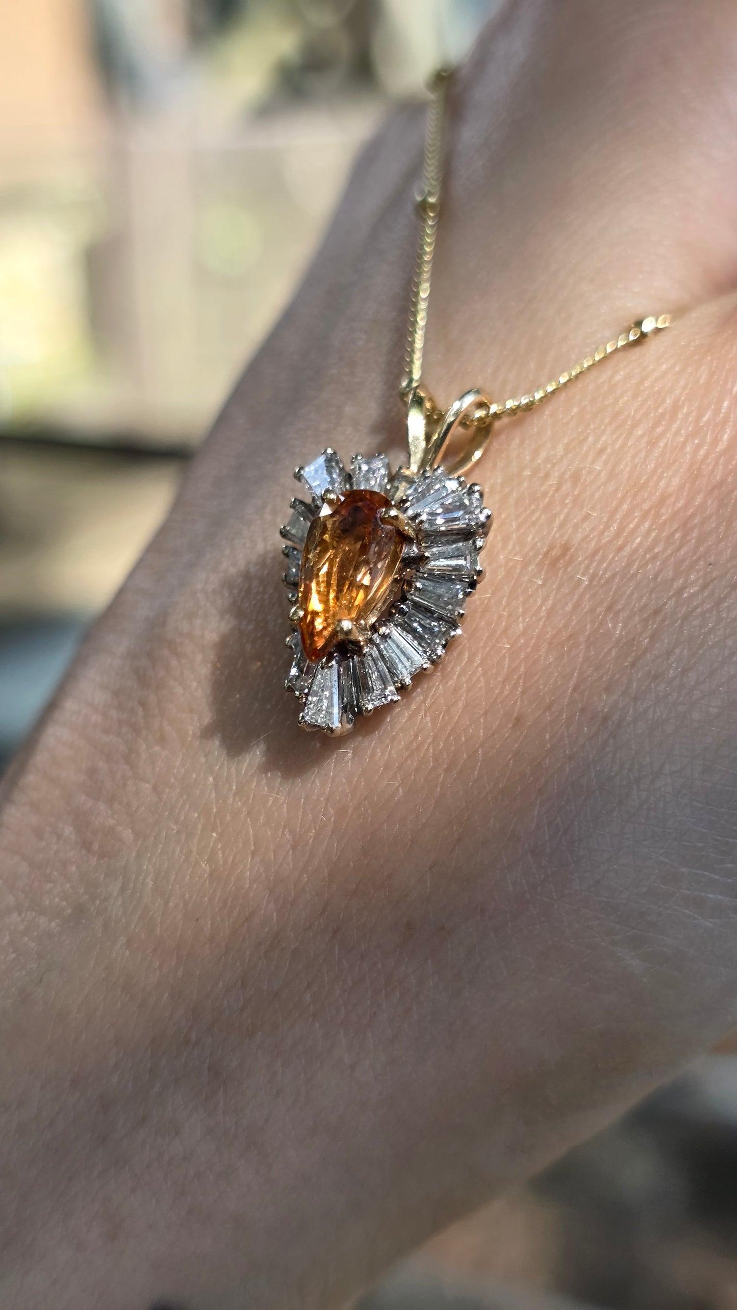 A gold pendant necklace with a marquise-cut amber gemstone surrounded by baguette-cut clear stones, shown worn on a person's wrist.