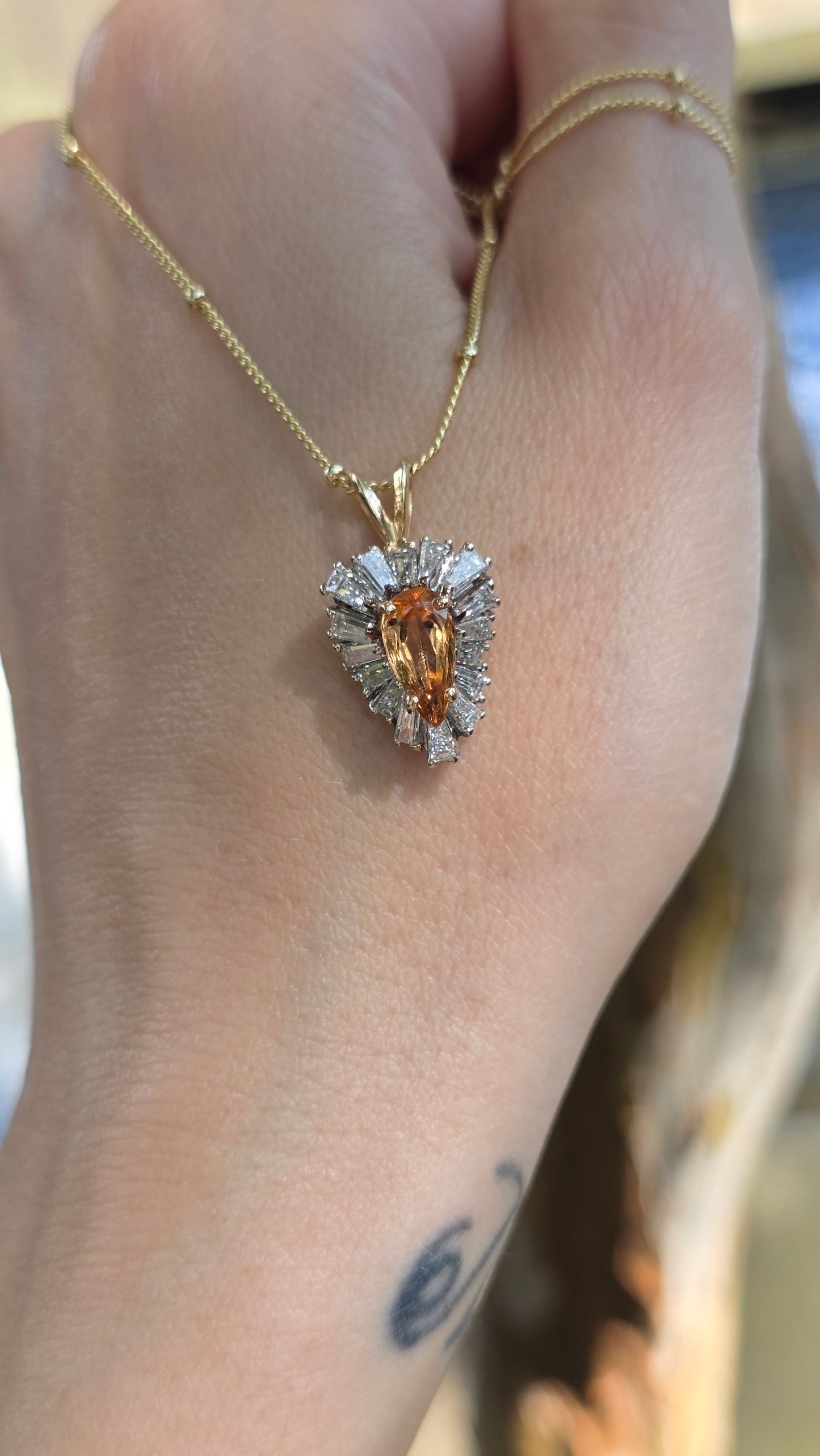 A gold chain necklace with a heart-shaped pendant featuring a central orange gemstone surrounded by clear stones, worn on a person's hand.