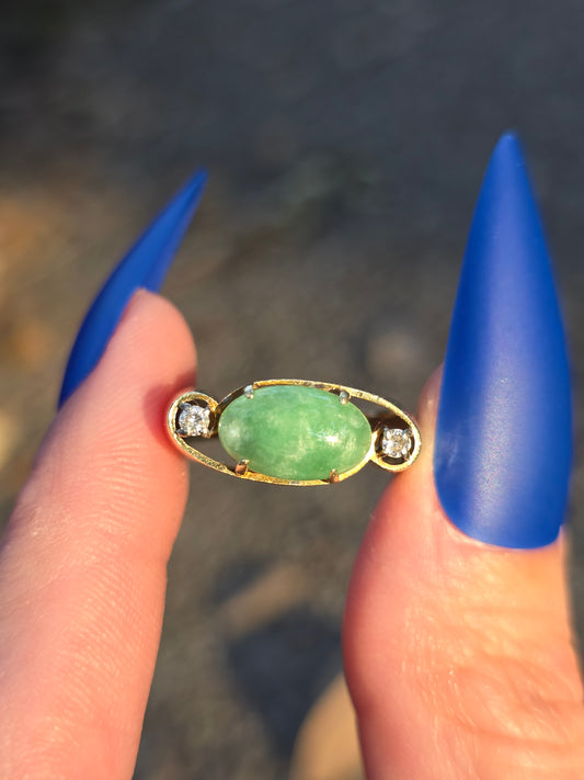 Green gemstone pendant with gold frame held between blue-tipped fingers against a blurred background