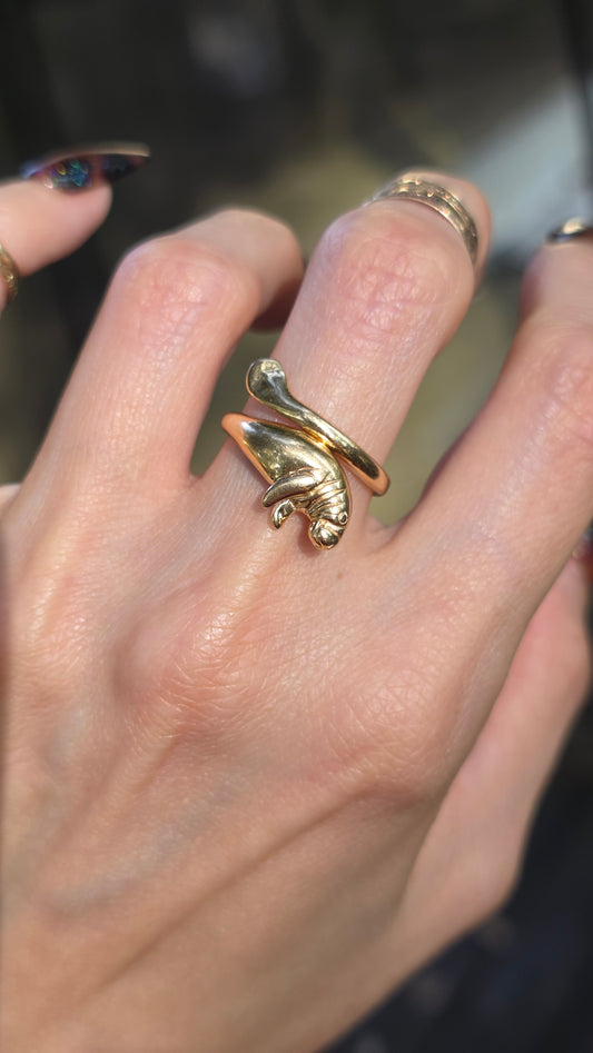 A close-up of a hand wearing a gold snake ring with a small manatee figure coiled on it, along with other rings on adjacent fingers.