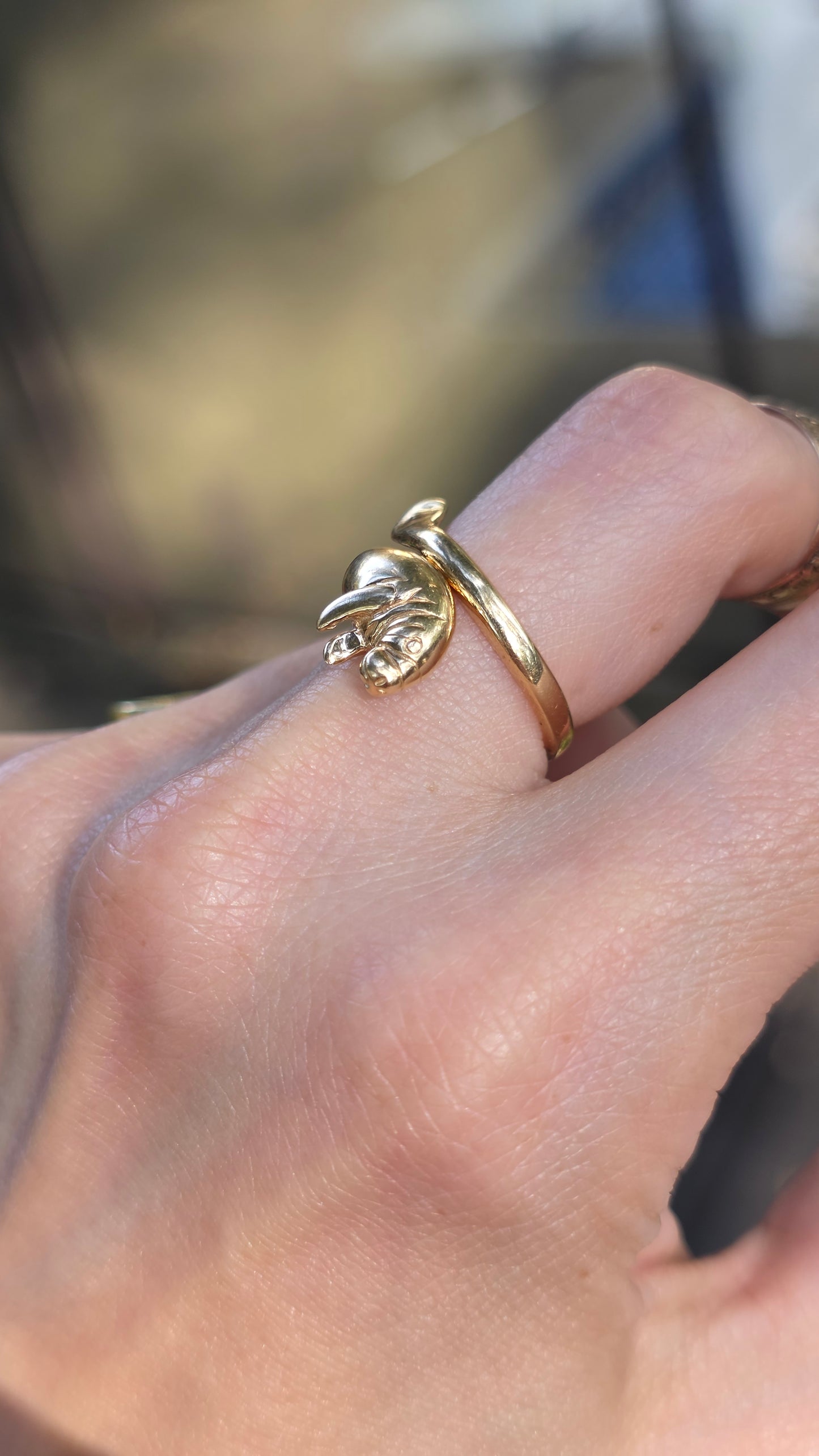 A close-up of a hand wearing a delicate gold ring with a small sculpted animal figure, likely a manatee, on the ring finger.
