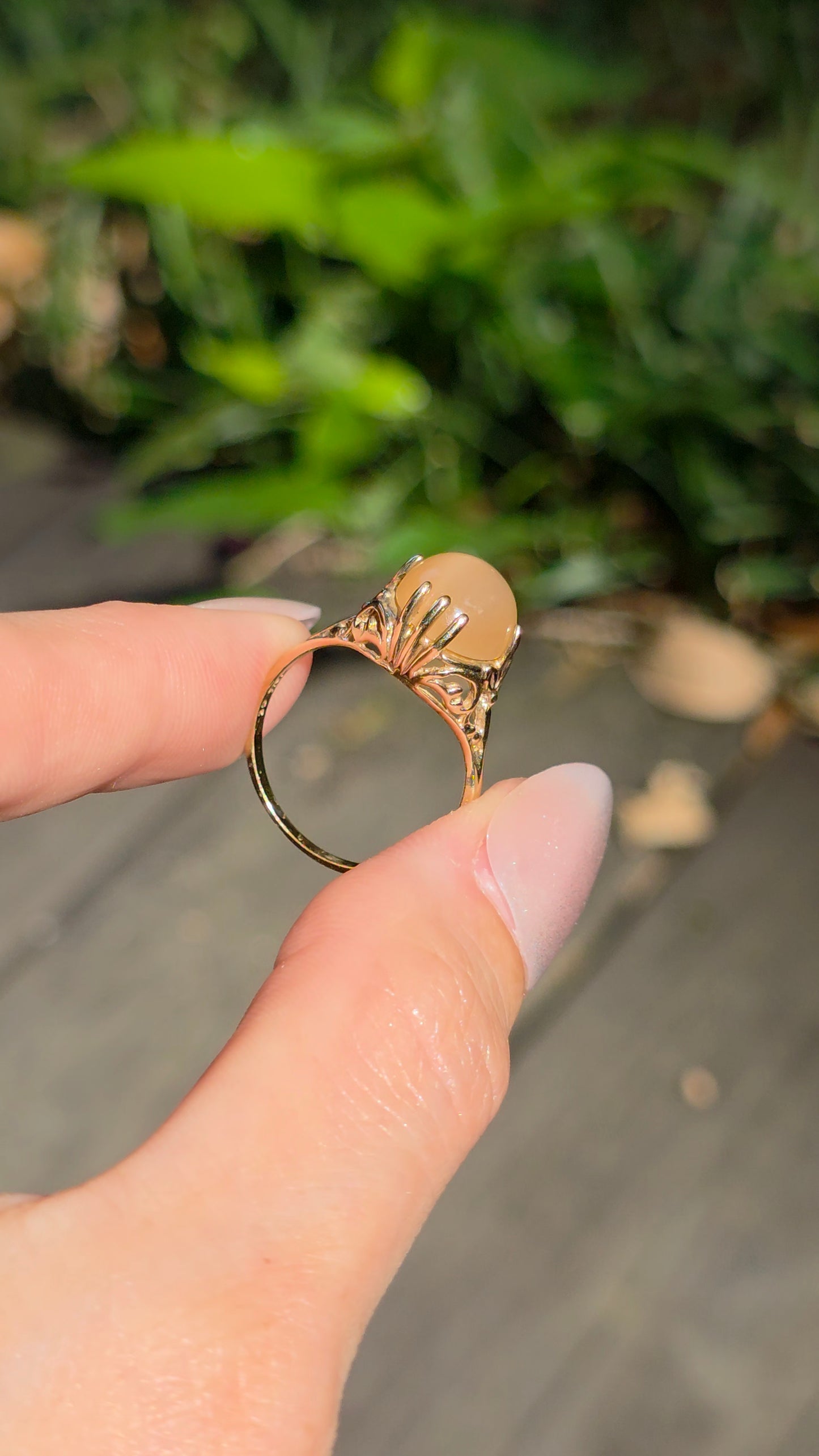 A hand holding a gold ring with a fan-shaped sunburst design and a central peach-colored stone, set against a blurred outdoor background of green foliage and a wooden deck.