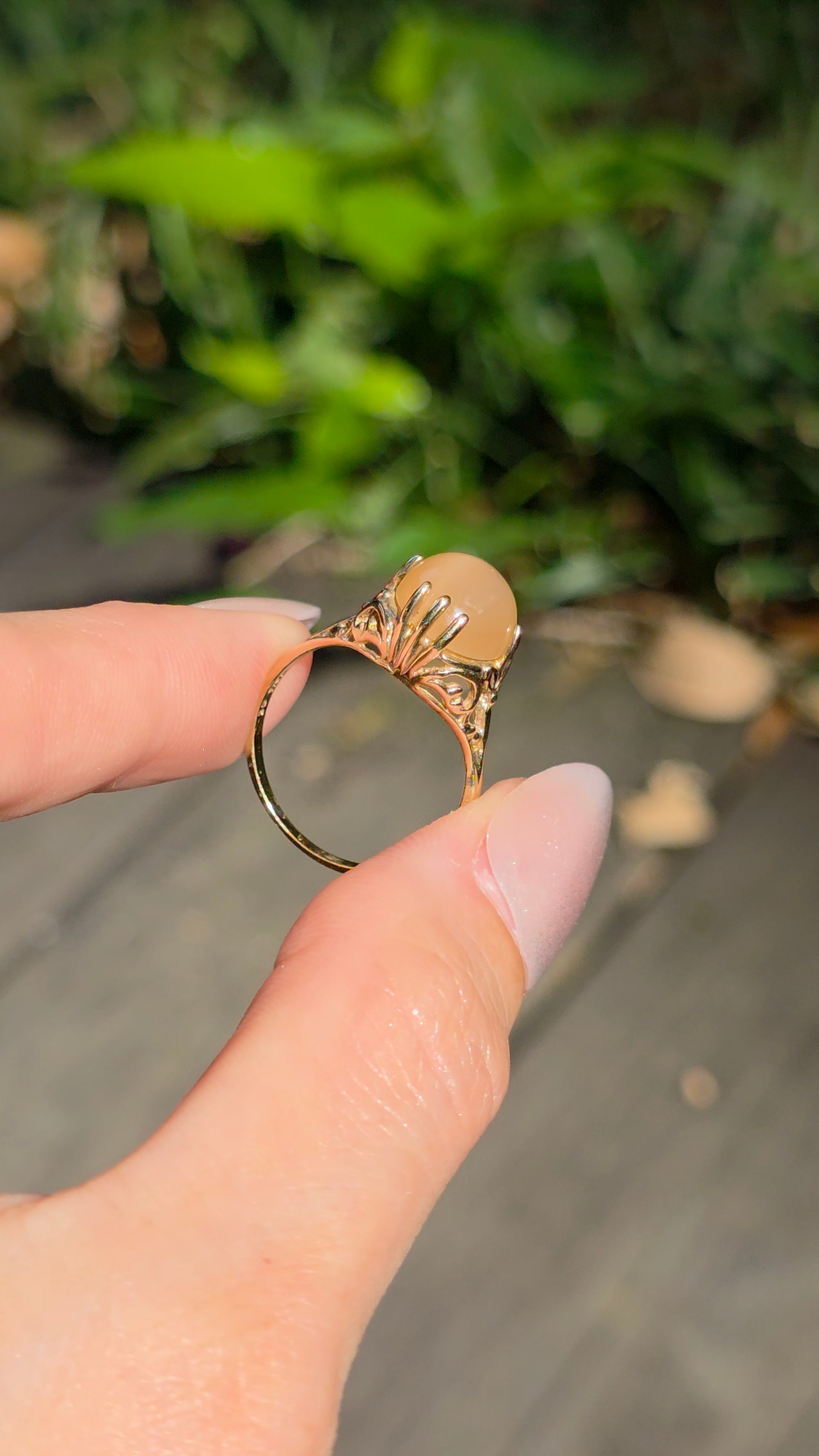 A hand holding a gold ring with a fan-shaped sunburst design and a central peach-colored stone, set against a blurred outdoor background of green foliage and a wooden deck.