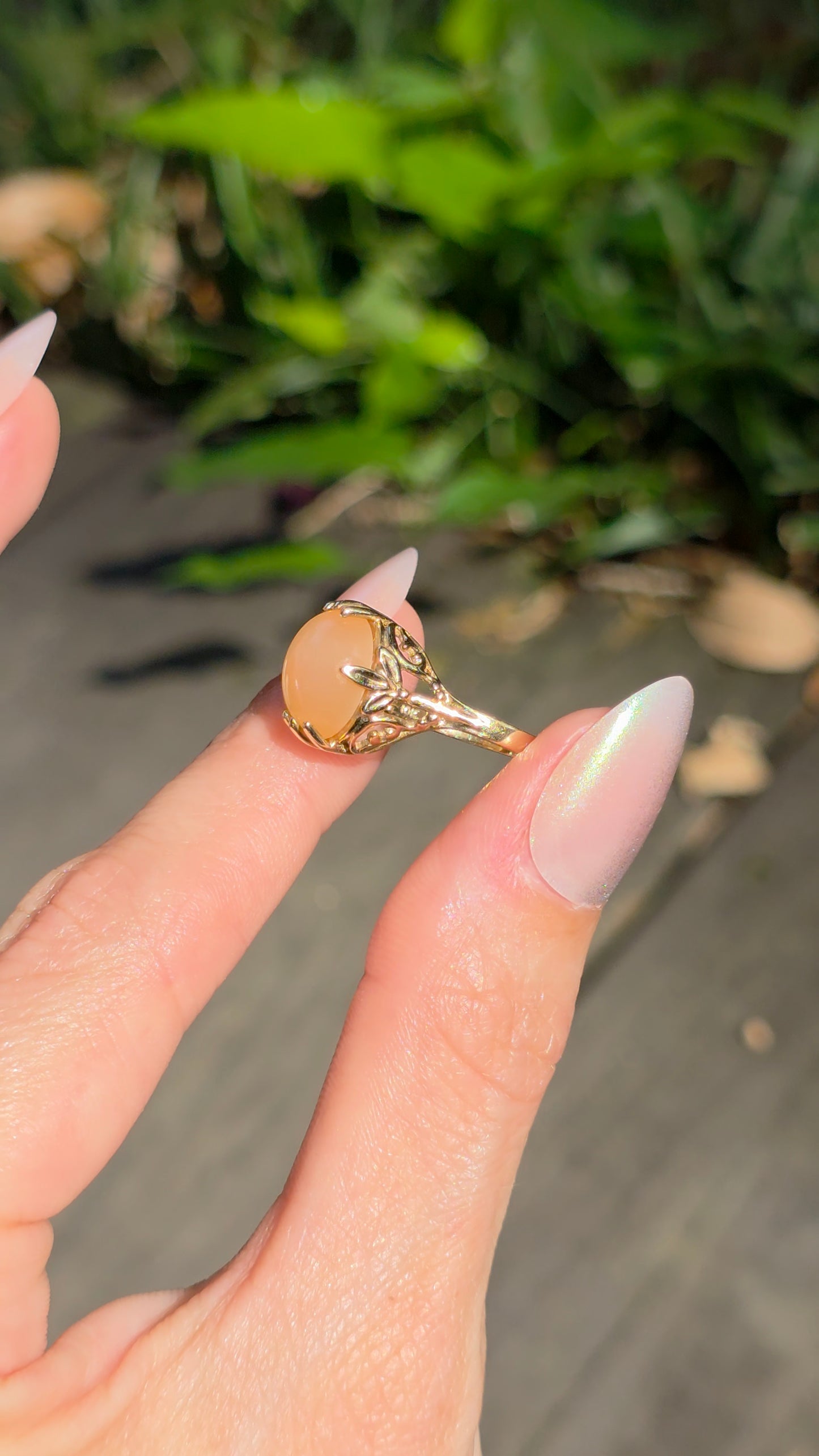 A gold ring with a large oval peach-colored stone held between fingers against a blurred outdoor background of green plants and gray pavement.