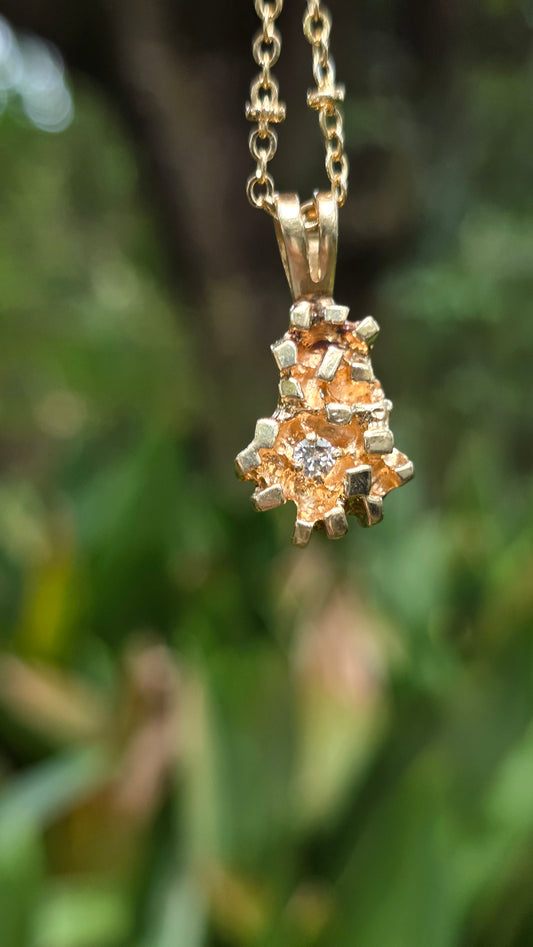 A gold pendant necklace featuring a cluster of small warm-toned stones with a central clear gemstone, hanging against a blurred green outdoor background.