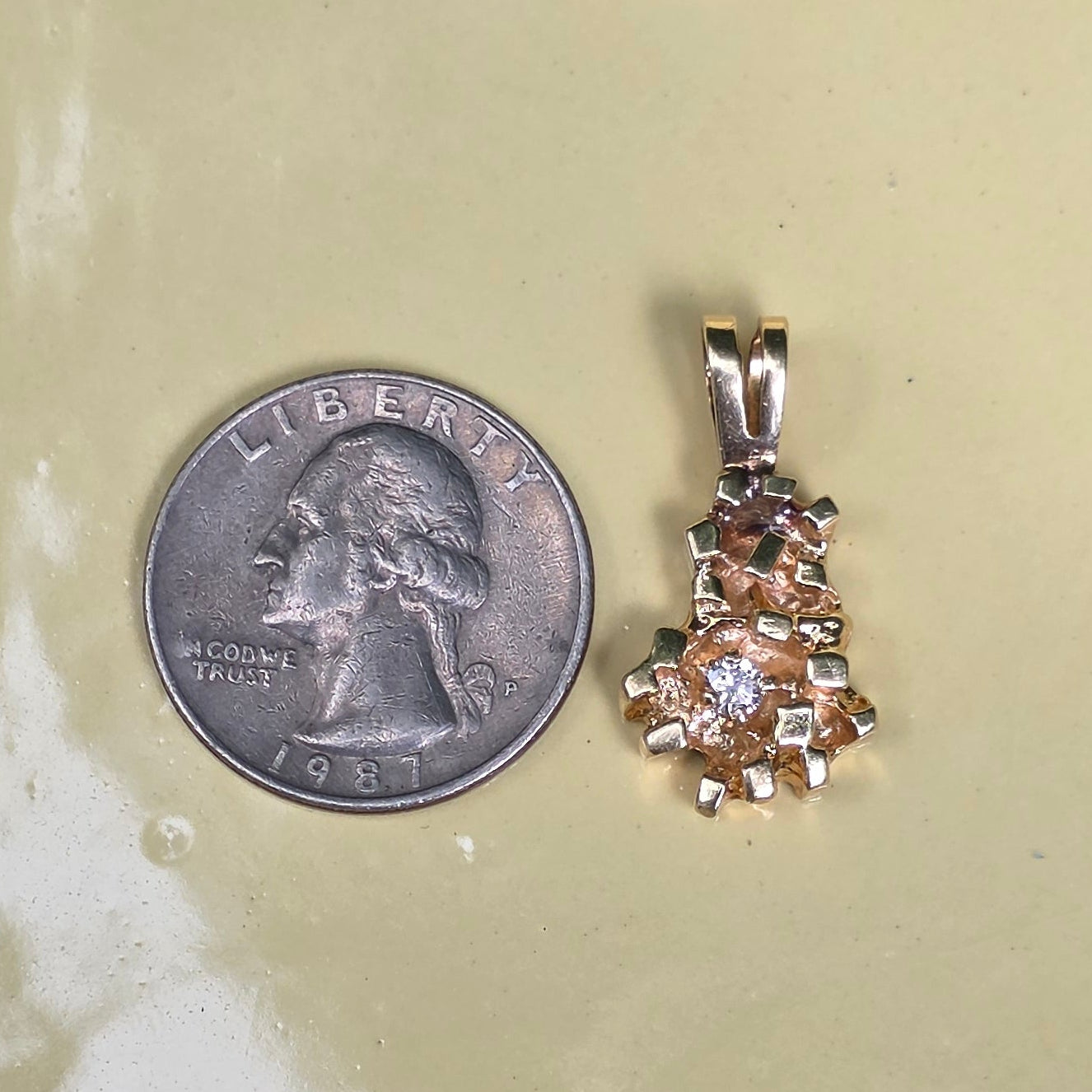 A small gold pendant with clear stones is displayed next to a US quarter coin on a pale yellow scalloped ceramic dish, set on a dark wood surface.