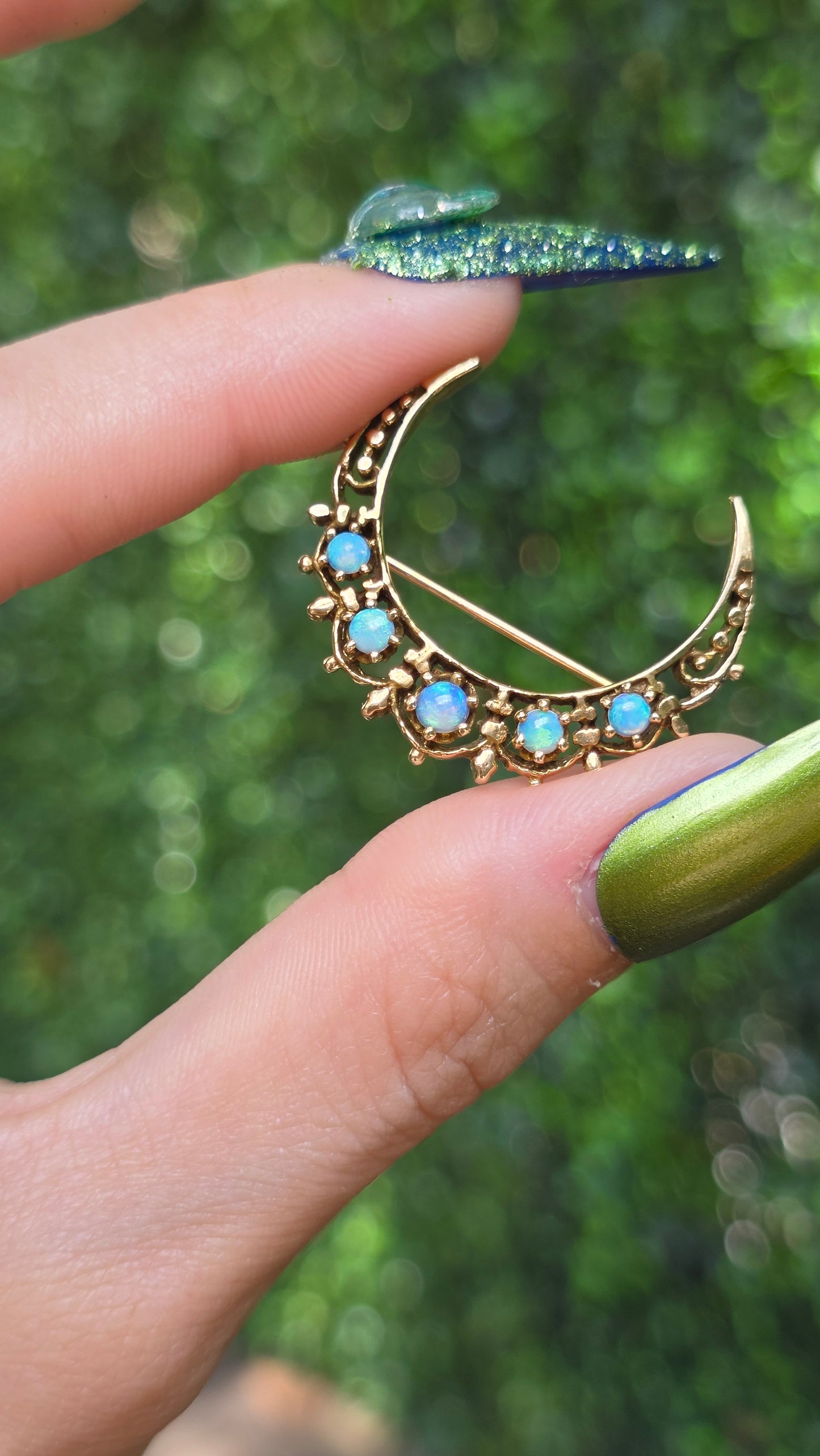 A gold crescent moon brooch with small light blue opal-like stones, held between fingers against a blurred green outdoor background.