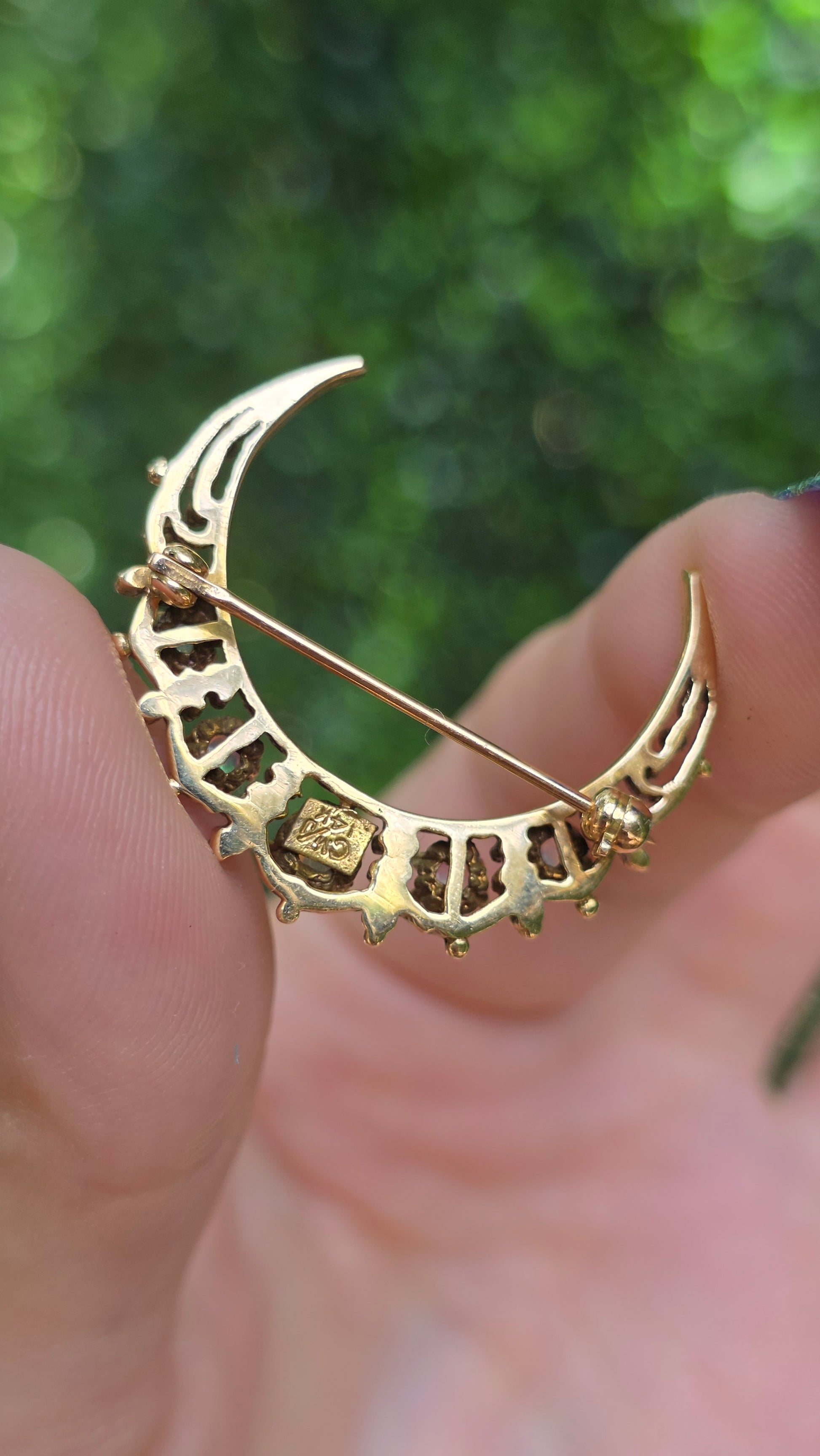 A gold crescent moon brooch with cutout details and a central square motif, held in a person's hand against a blurred green outdoor background.