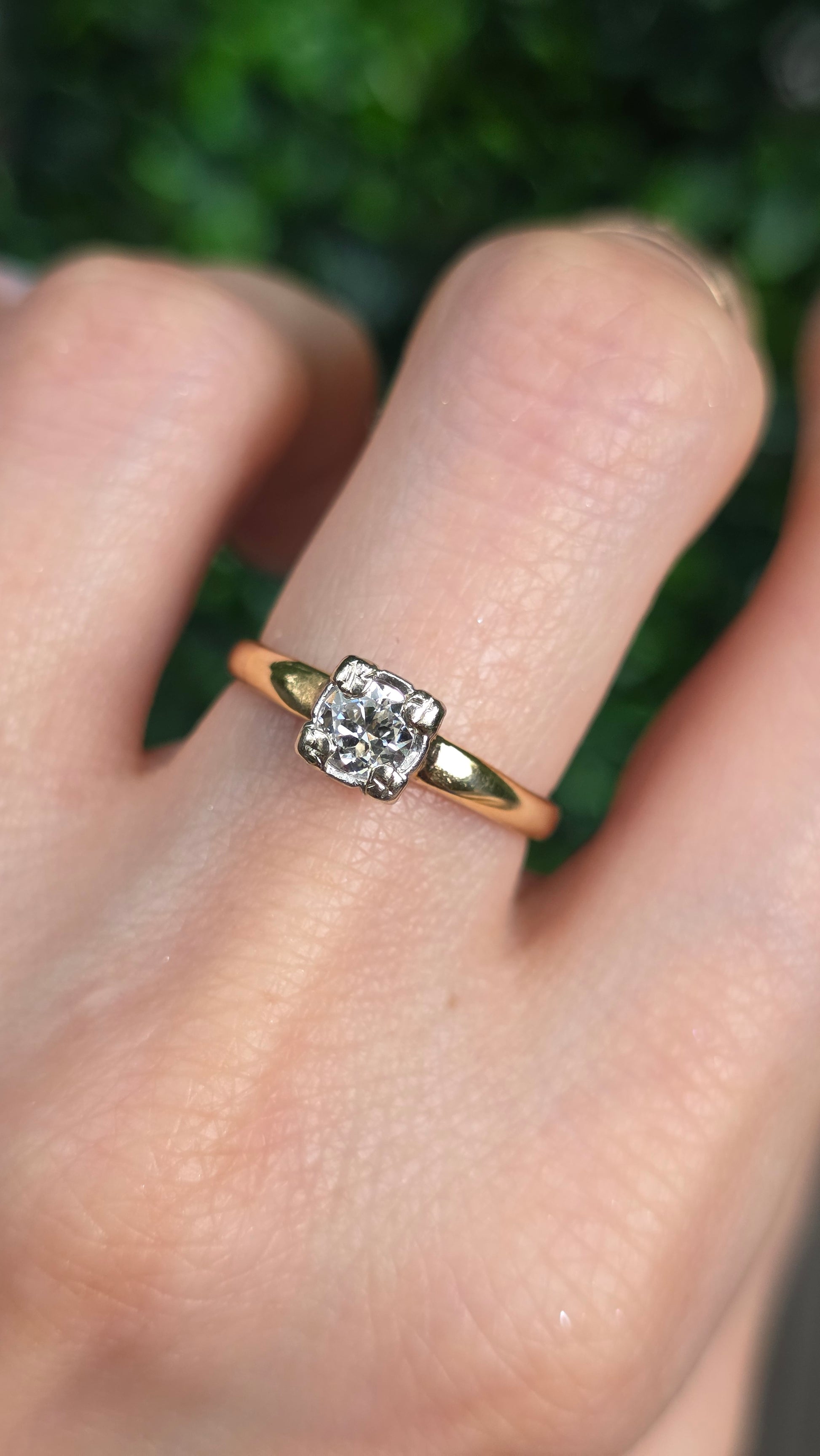 A close-up of a hand wearing a yellow gold ring with a square-cut clear gemstone in a four-prong setting, with a blurred green background.