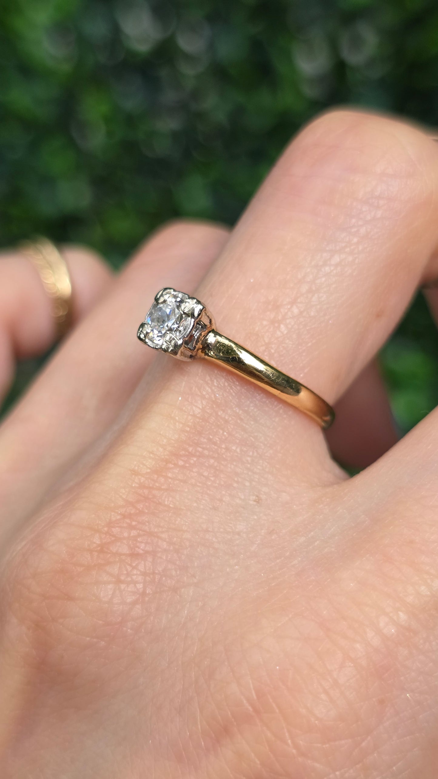 A close-up of a hand wearing a yellow gold ring with a square-cut clear gemstone, set against a blurred green outdoor background.