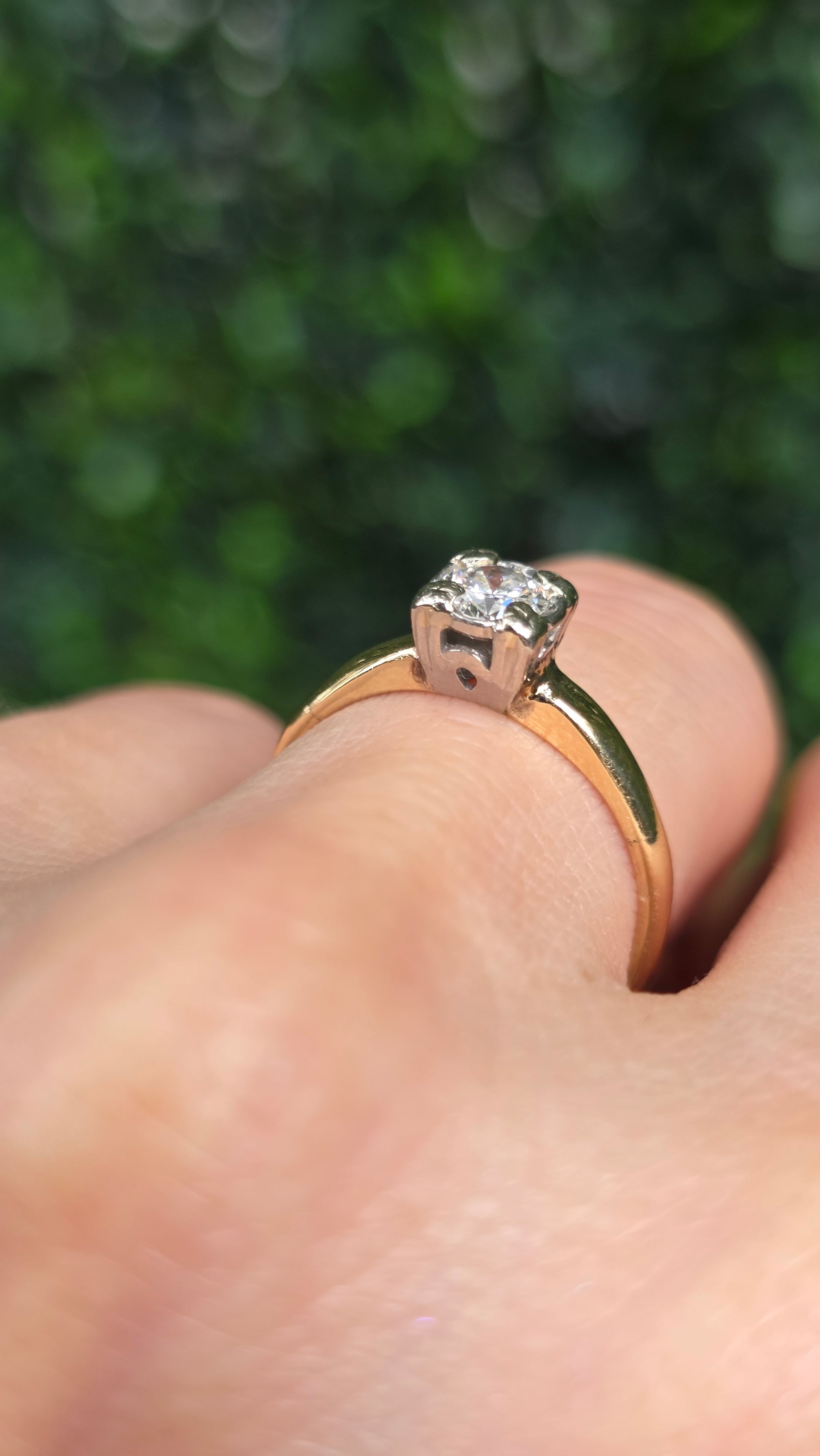 A close-up of a gold ring with a clear gemstone worn on a finger, set against a blurred green outdoor background.