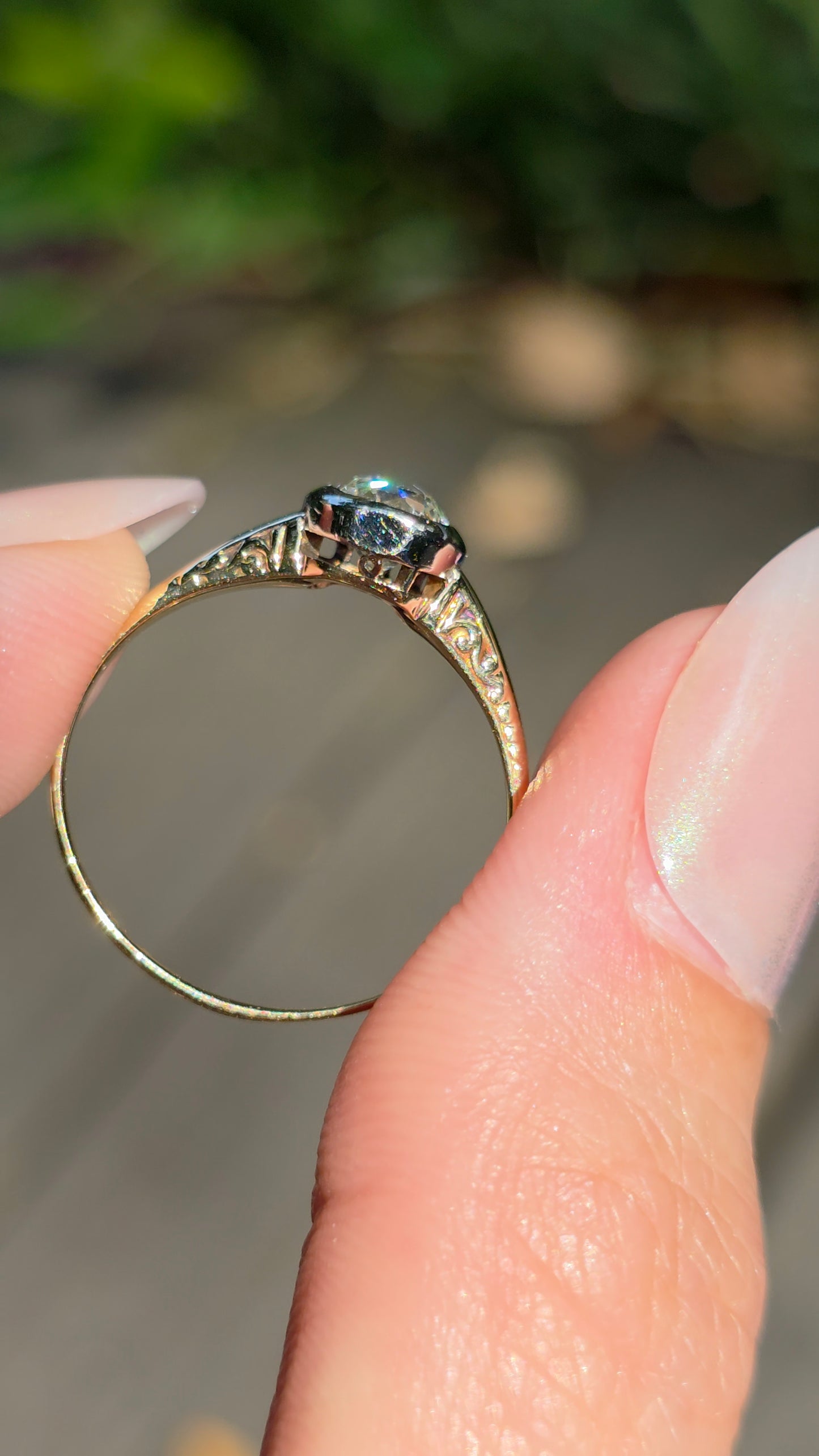 A close-up of a hand holding a yellow gold ring with a dark, possibly black or oxidized, decorative setting and a small central gemstone, against a blurred outdoor background.