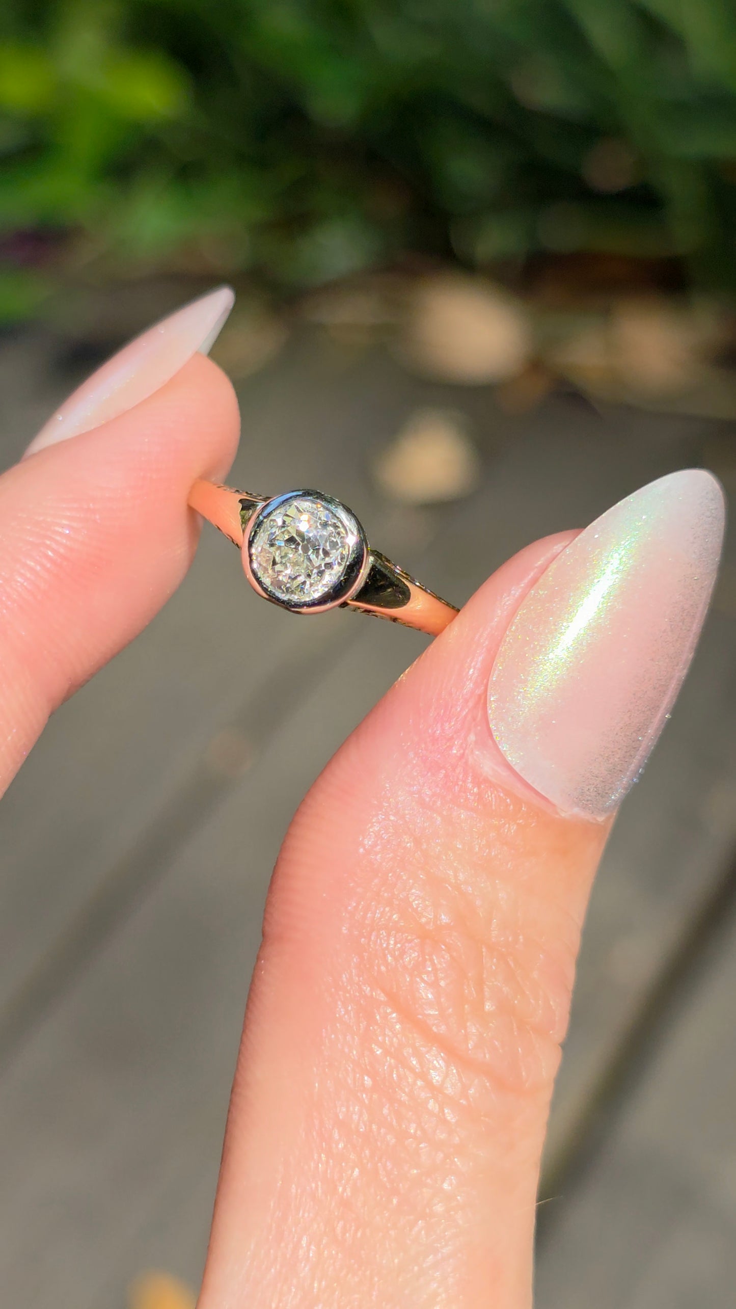 A close-up of a hand with long manicured nails holding a gold ring with a round clear gemstone, set against a blurred outdoor background.