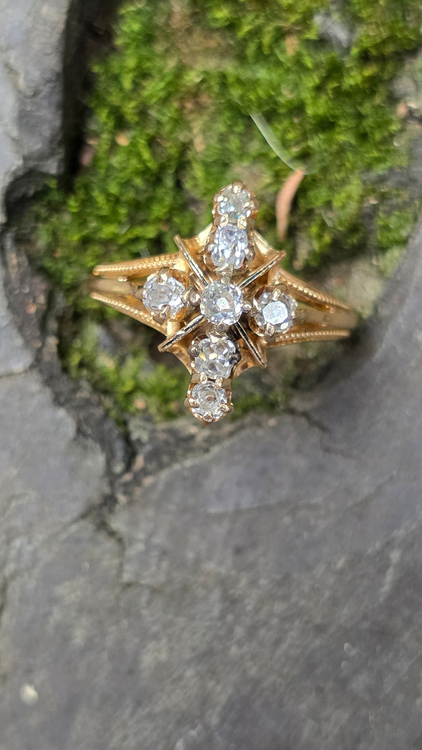 A gold ring with seven clear round-cut gemstones arranged in a floral or starburst pattern, resting on a mossy green stone surface against a gray stone background.