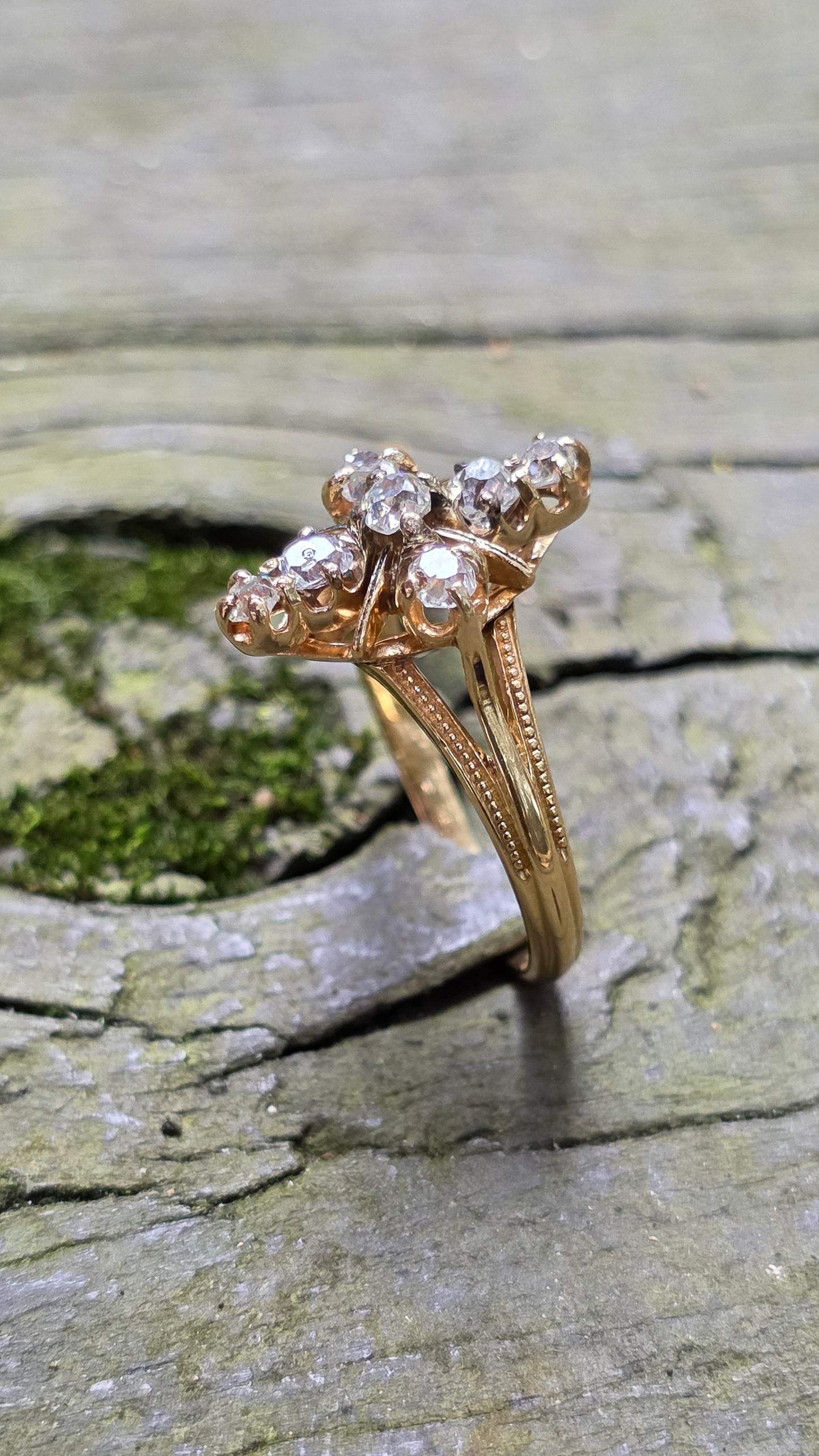 A vintage-style gold ring with a cluster of clear gemstones, photographed resting against a textured, mossy wood surface.