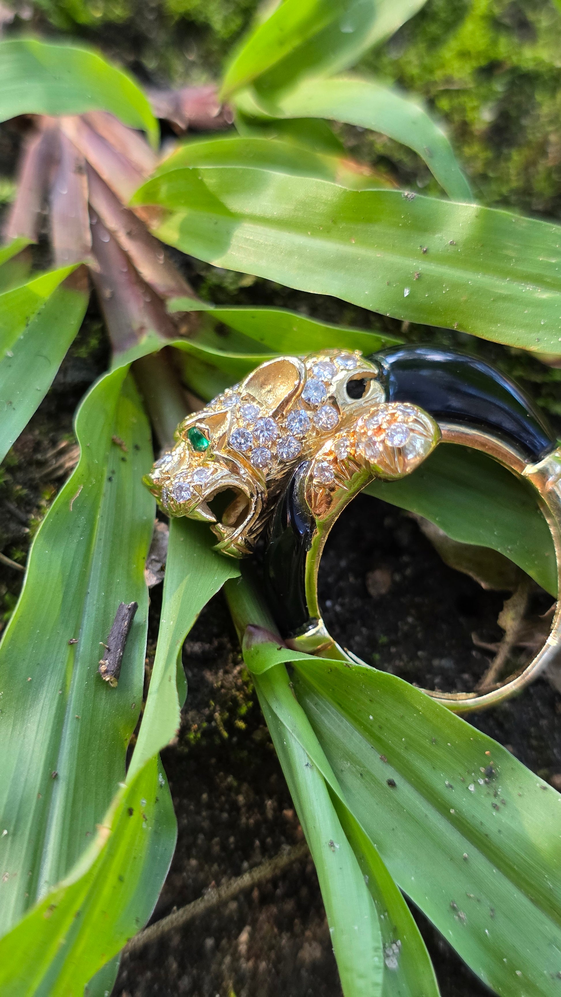 A gold-toned ring shaped like a panther head, decorated with pavé-set clear stones and a small green gemstone in the eye, resting on green leaves and dark soil.