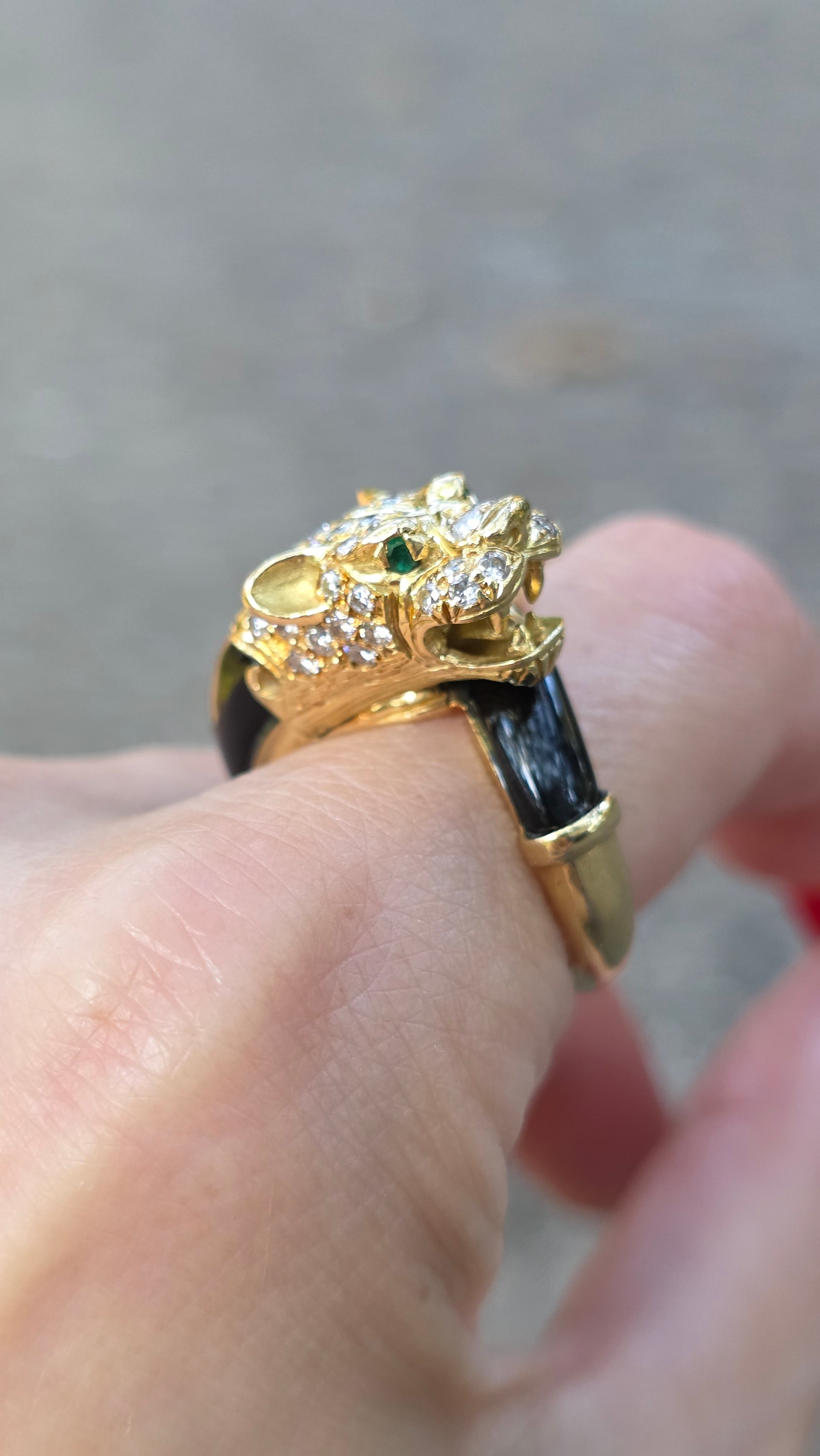 A close-up of a hand wearing a gold ring shaped like a lion head, decorated with small clear stones and green gem accents, with a black band.
