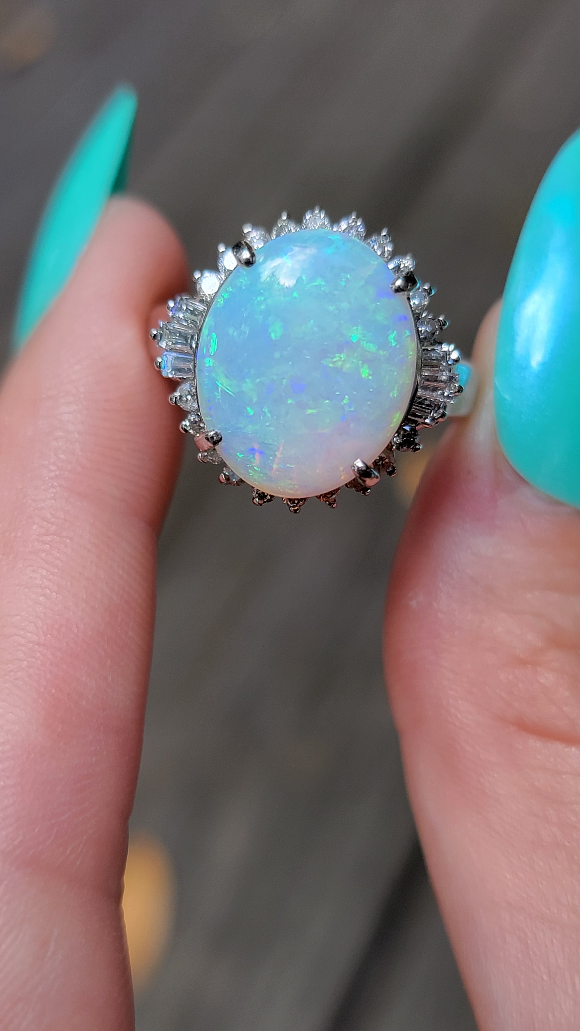 A vintage platinum ring with a large opal center stone and smaller diamond surrounding stones, held between fingers with teal nail polish.
