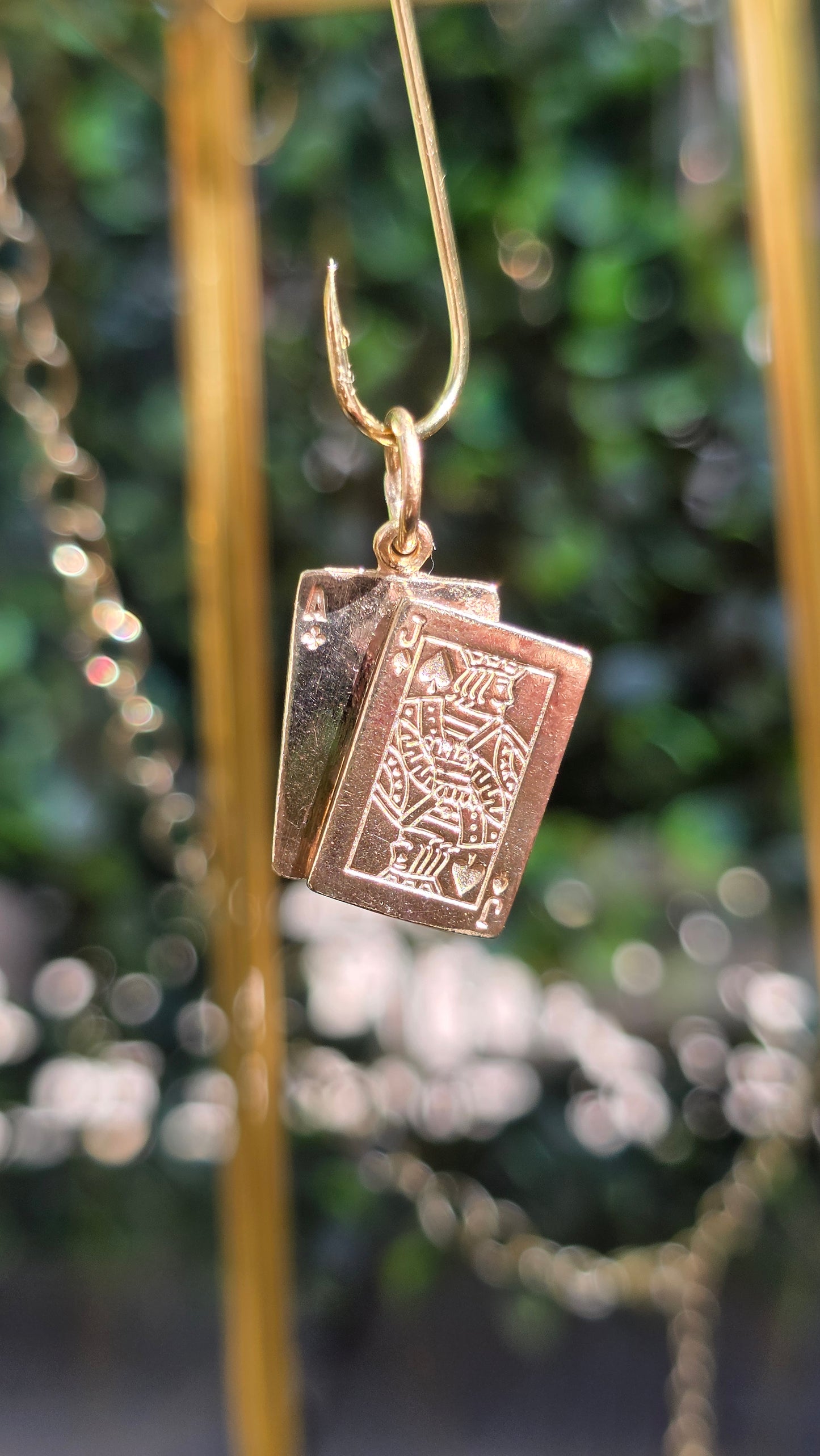 A close-up of a gold-toned earring with a rectangular engraved pendant, hanging against a blurred green and white bokeh background.