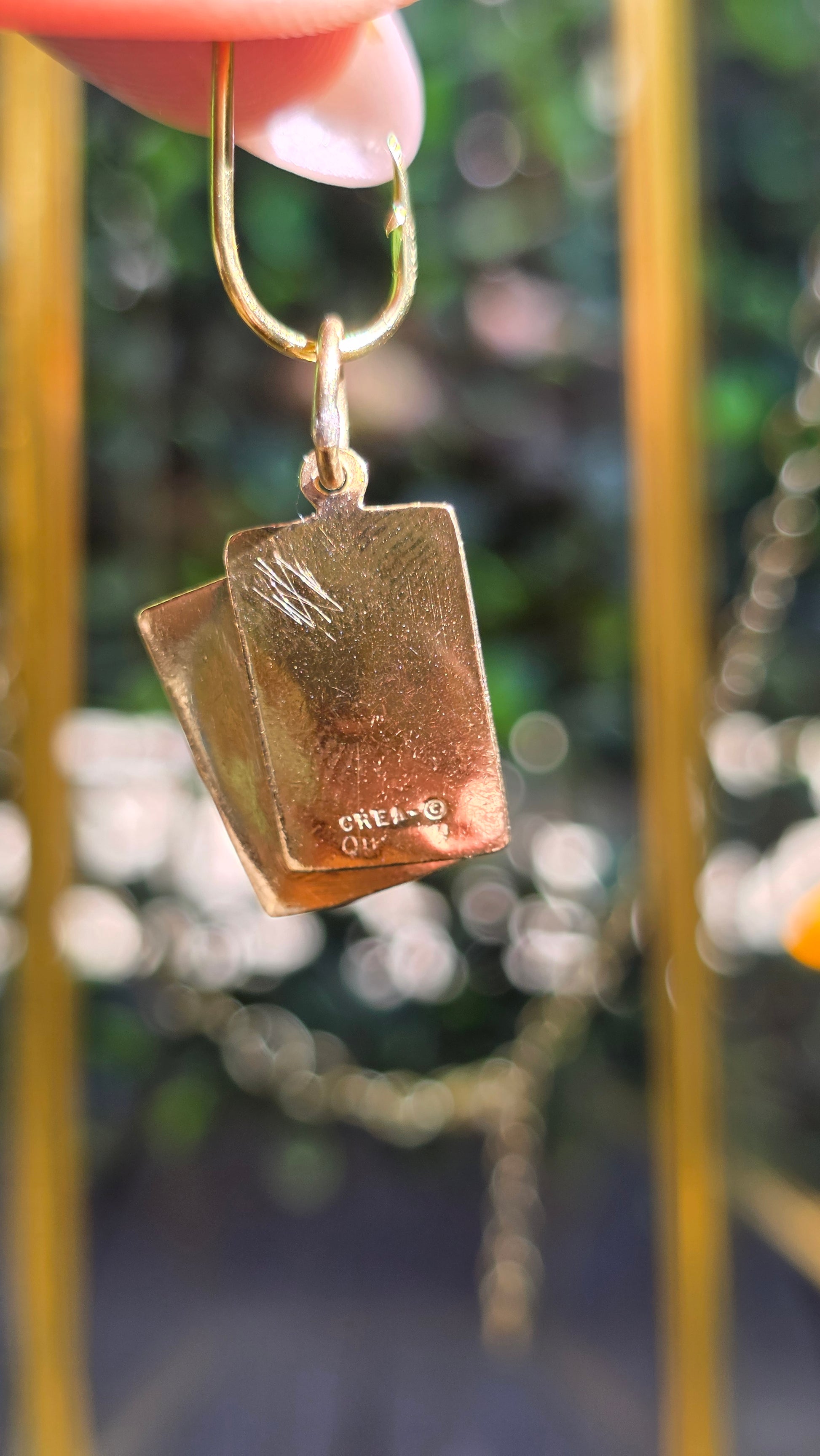 A close-up of a hand holding a gold earring with a square, textured gold pendant that has a small engraved 'GIVREAU' logo, set against a blurred background.