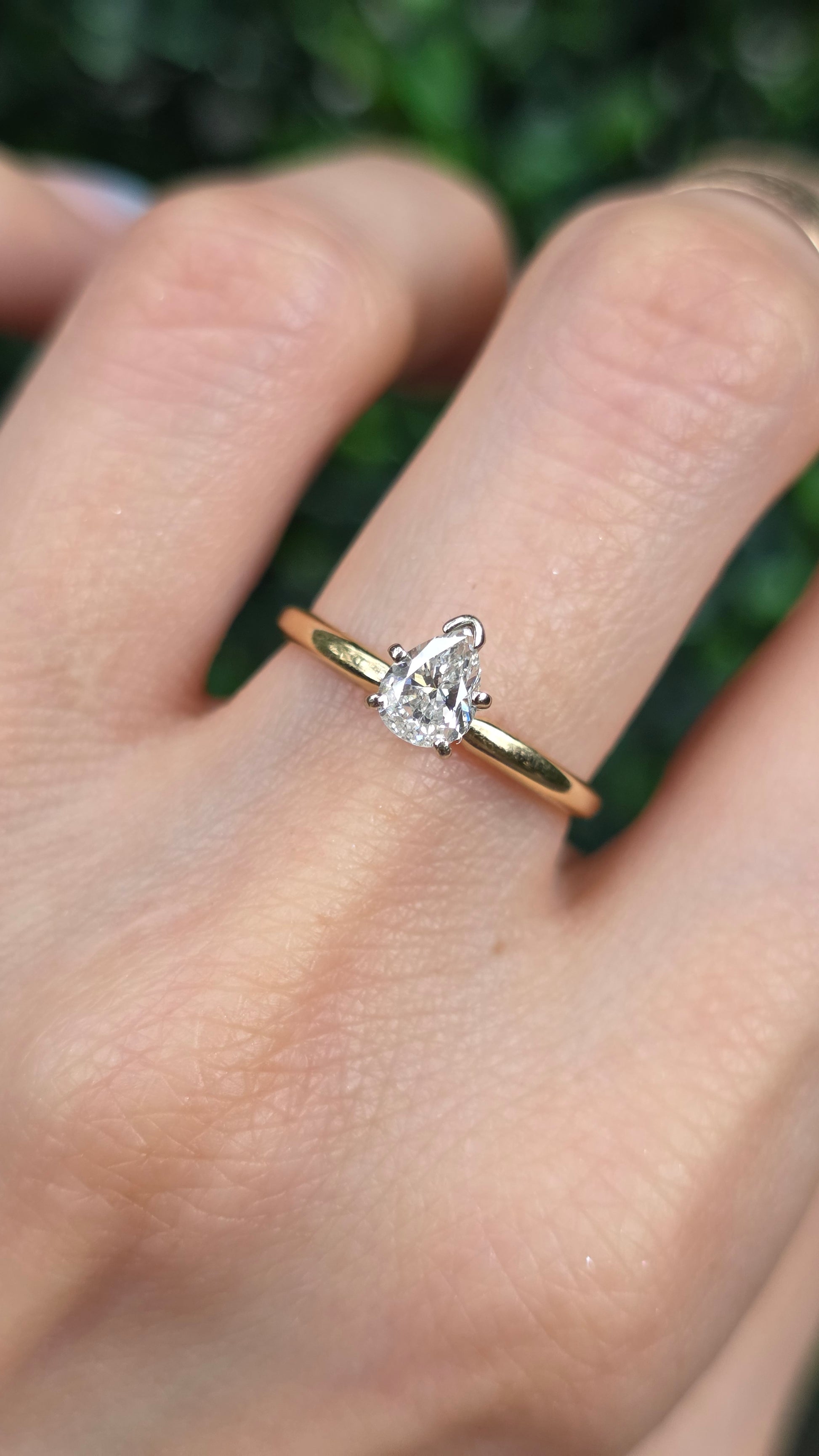 A close-up of a hand wearing a gold ring with a pear-shaped clear gemstone, set against a blurred green background.