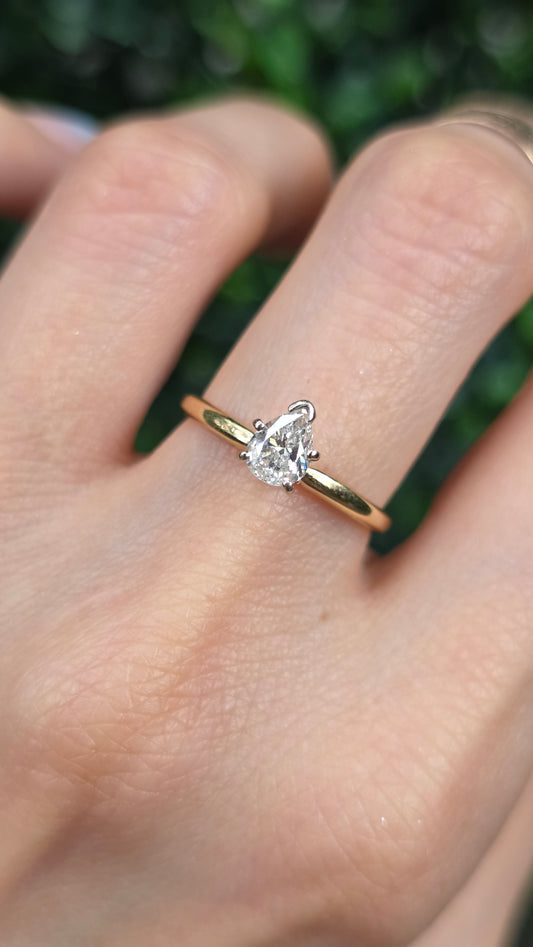 A close-up of a hand wearing a gold ring with a pear-shaped clear gemstone, set against a blurred green background.