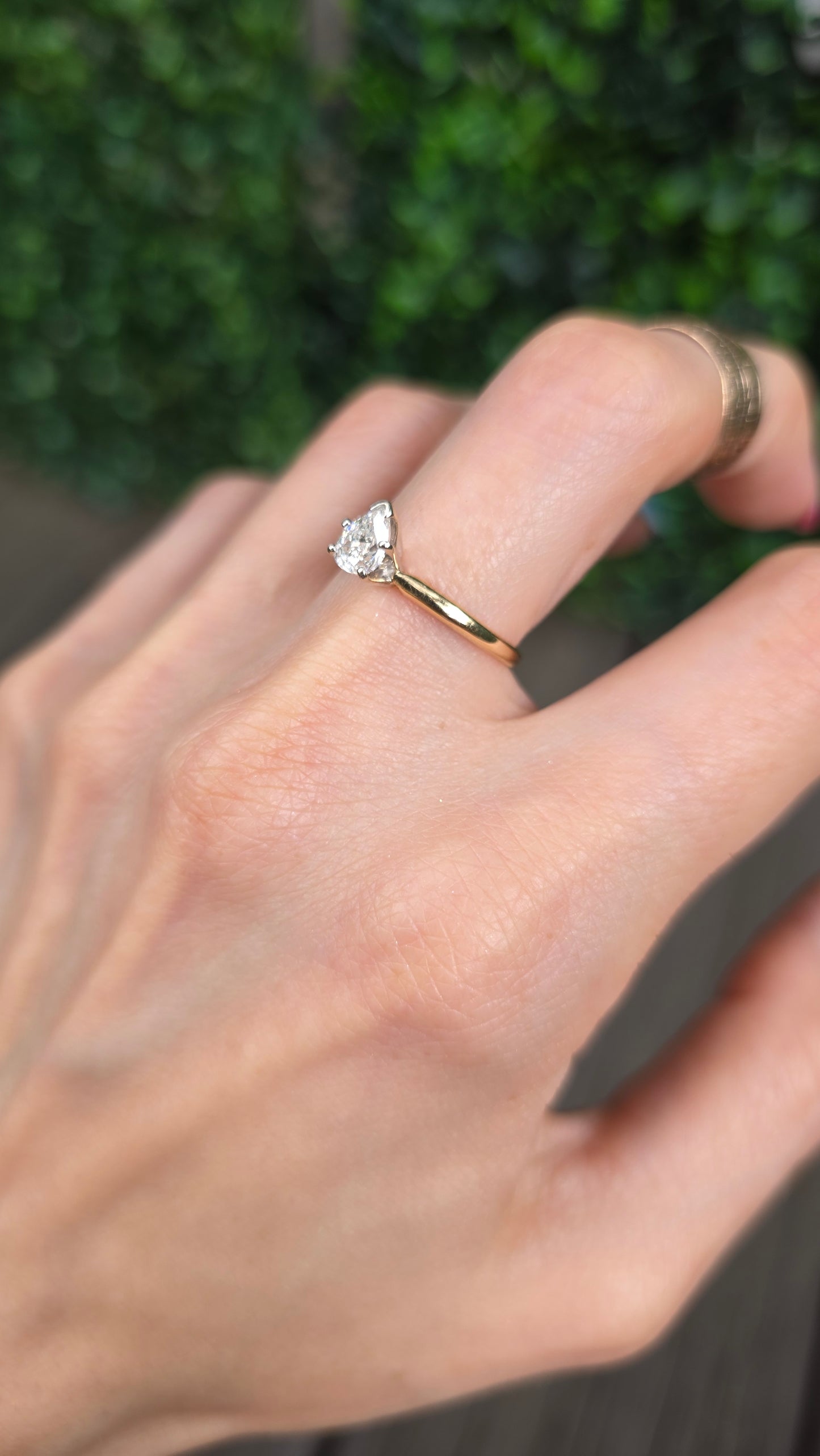 A close-up of a hand wearing a delicate gold ring with a small, irregular-cut clear gemstone, set against a blurred green background.