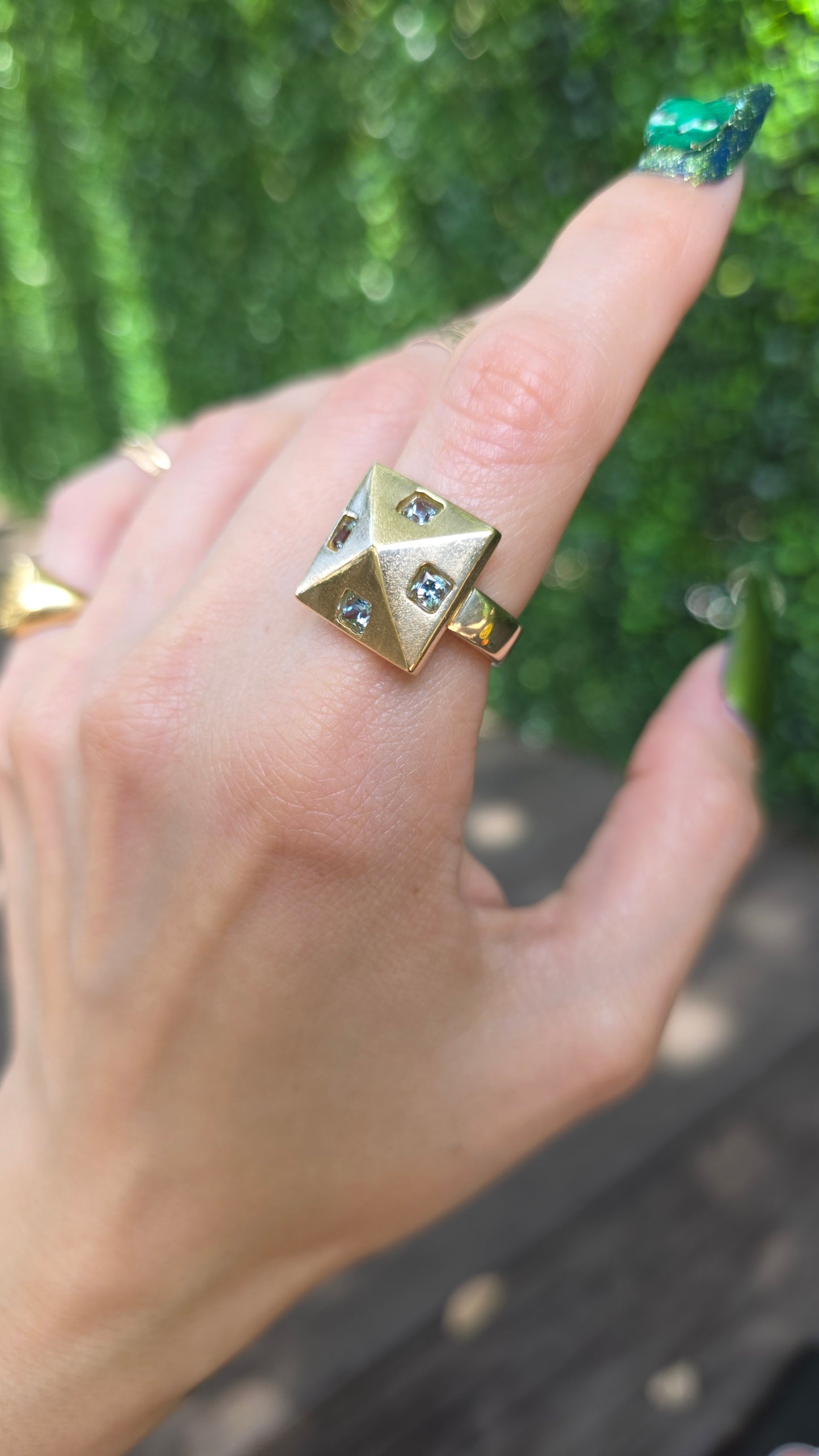 A gold pyramid ring with small clear gemstones on the fingers of a hand, photographed outdoors with a blurred green and brown background.