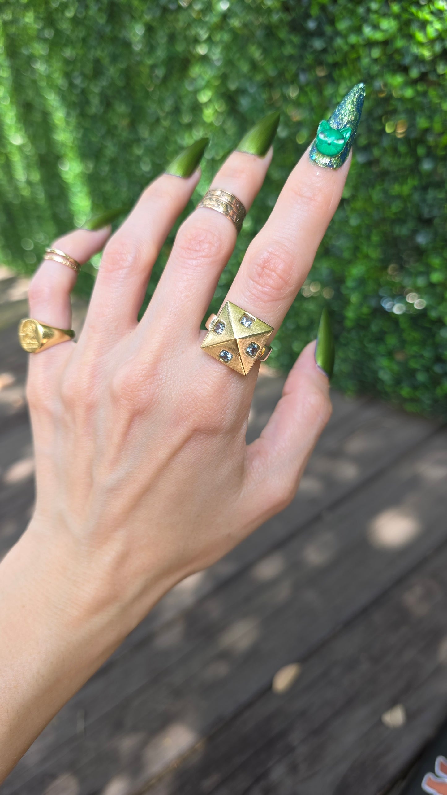 A close-up of a hand wearing multiple rings, including a prominent gold ring with a triangular cutout design and small dark stones, set against a blurred background of green foliage and a wooden deck.