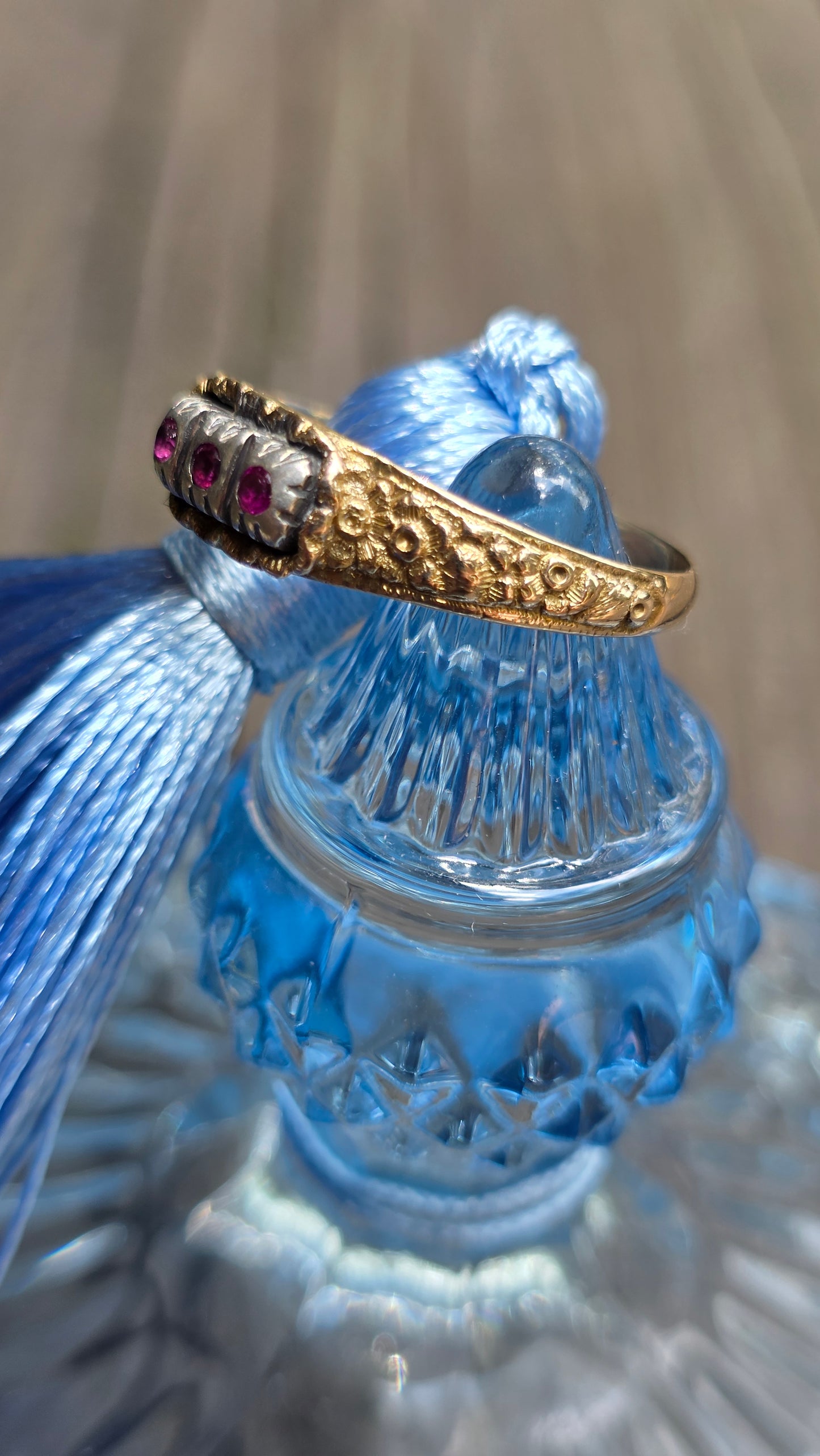 A gold ring with a floral crown design and small pink gemstones, displayed on a blue glass tassel holder.