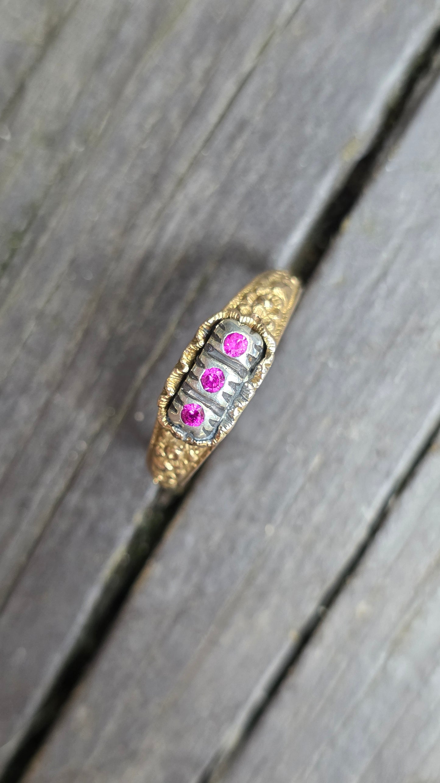 A vintage-style gold ring with a silver-toned center band, featuring three small pink gemstones, resting on weathered gray wooden planks.