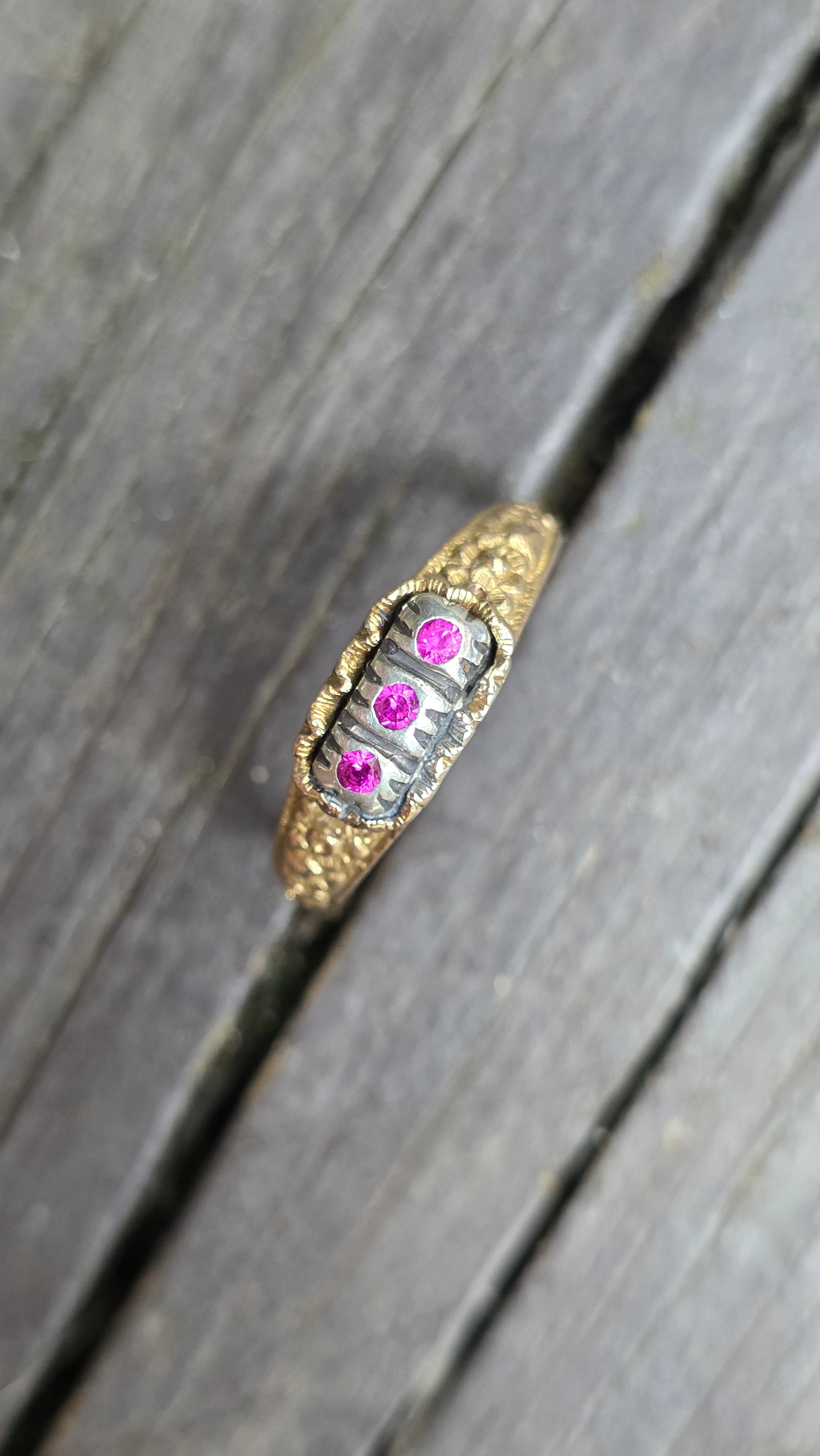 A vintage-style gold ring with a silver-toned center band, featuring three small pink gemstones, resting on weathered gray wooden planks.