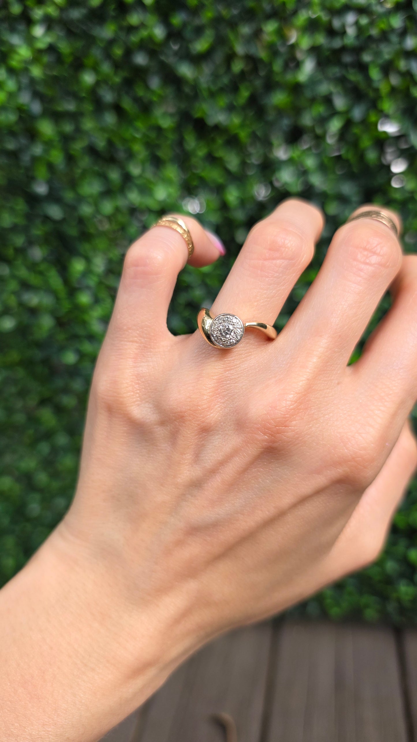 A close-up of a hand wearing a gold ring with a round clear gemstone, set against a blurred background of green foliage and a wooden surface.