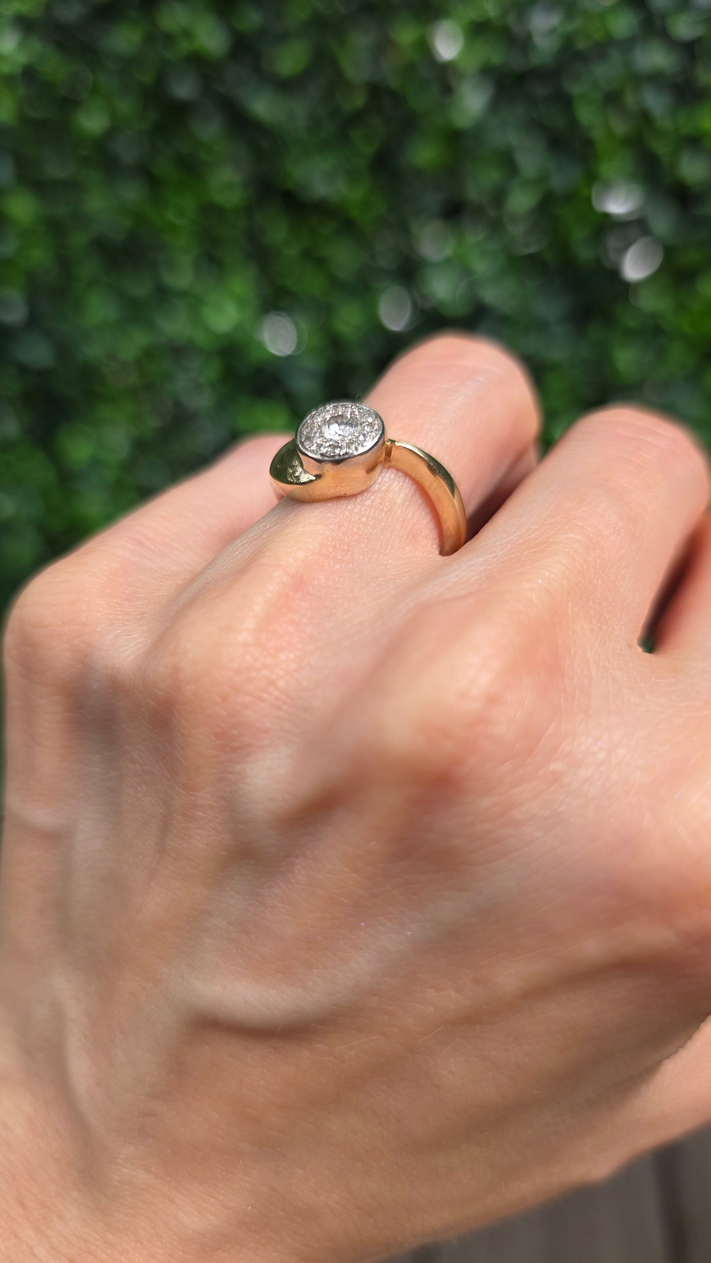 A close-up of a hand wearing a gold signet ring with a round clear gemstone, set against a blurred green background.