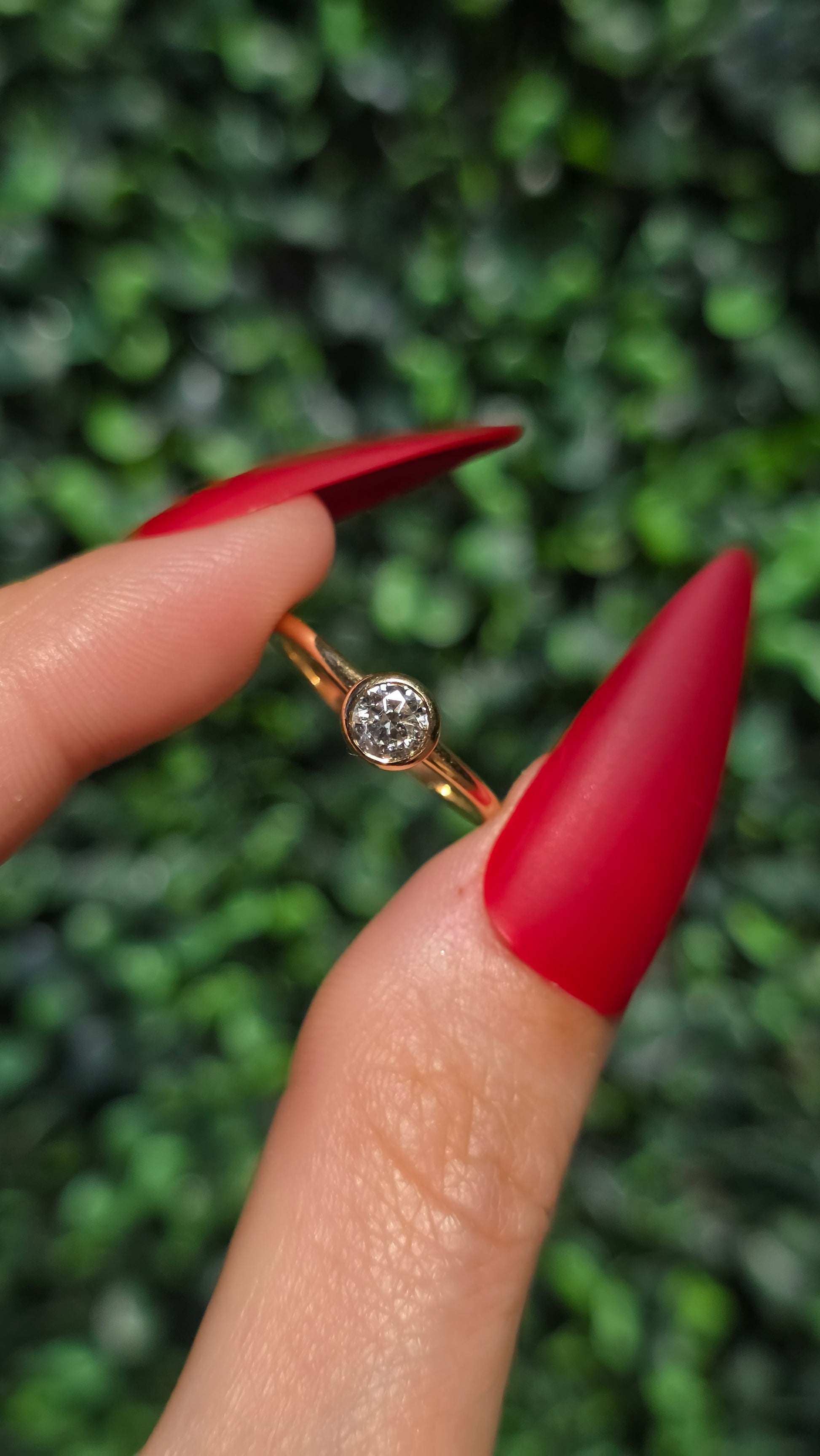 A close-up of a hand with long red manicured nails holding a delicate gold ring with a small clear gemstone, set against a blurred green foliage background.