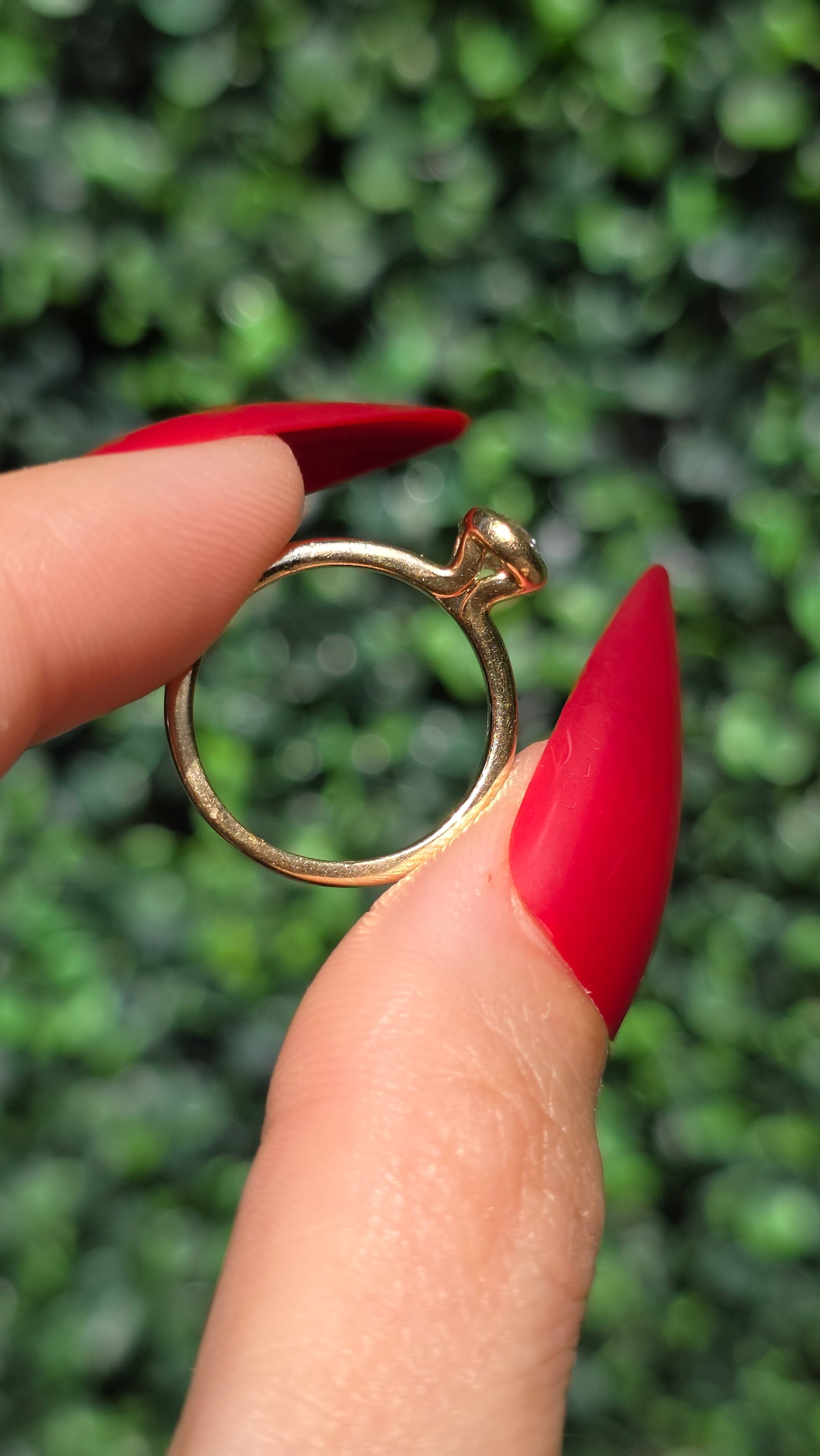 A gold ring with a small, dome-shaped clear gemstone held between two fingers with long, bright red manicured nails, set against a blurred green hedge background.