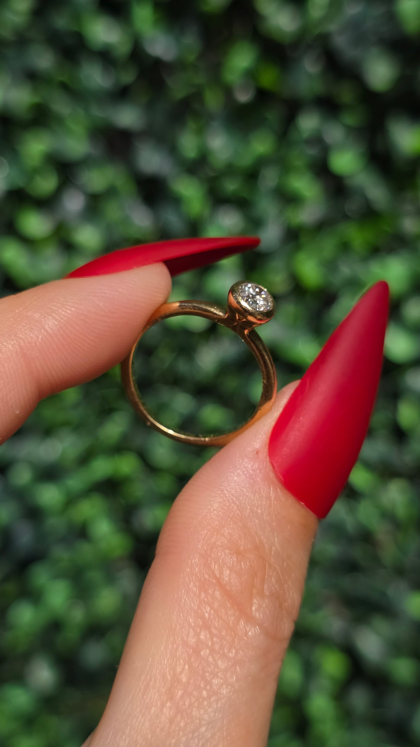 A gold ring with a single round-cut clear gemstone held between two fingers with long red manicured nails, set against a blurred green foliage background.