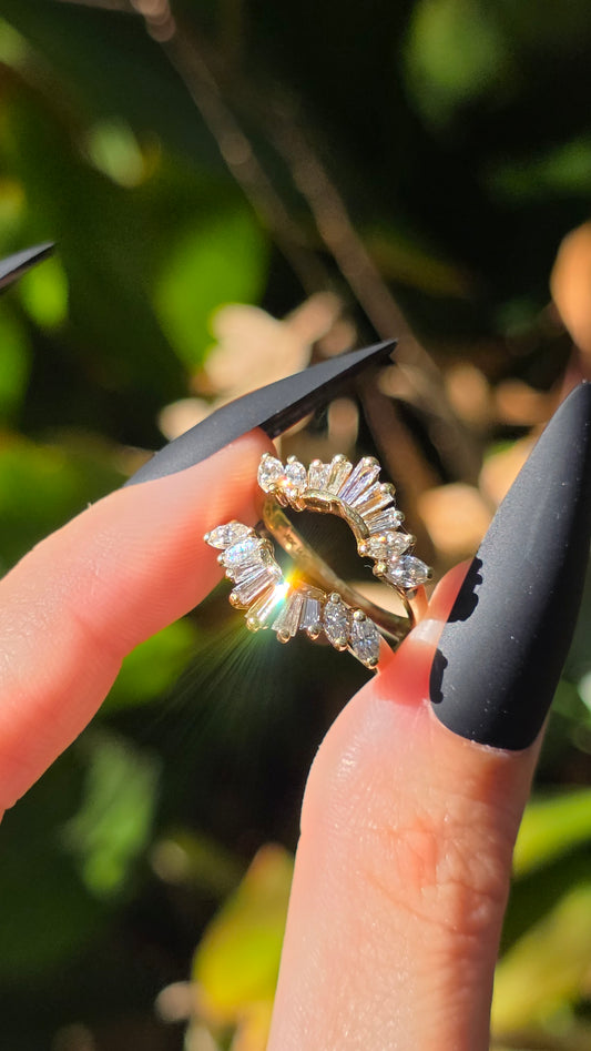 A gold ring with a halo of clear baguette and round-cut stones, held between two fingers with long black manicured nails against a blurred green outdoor background.