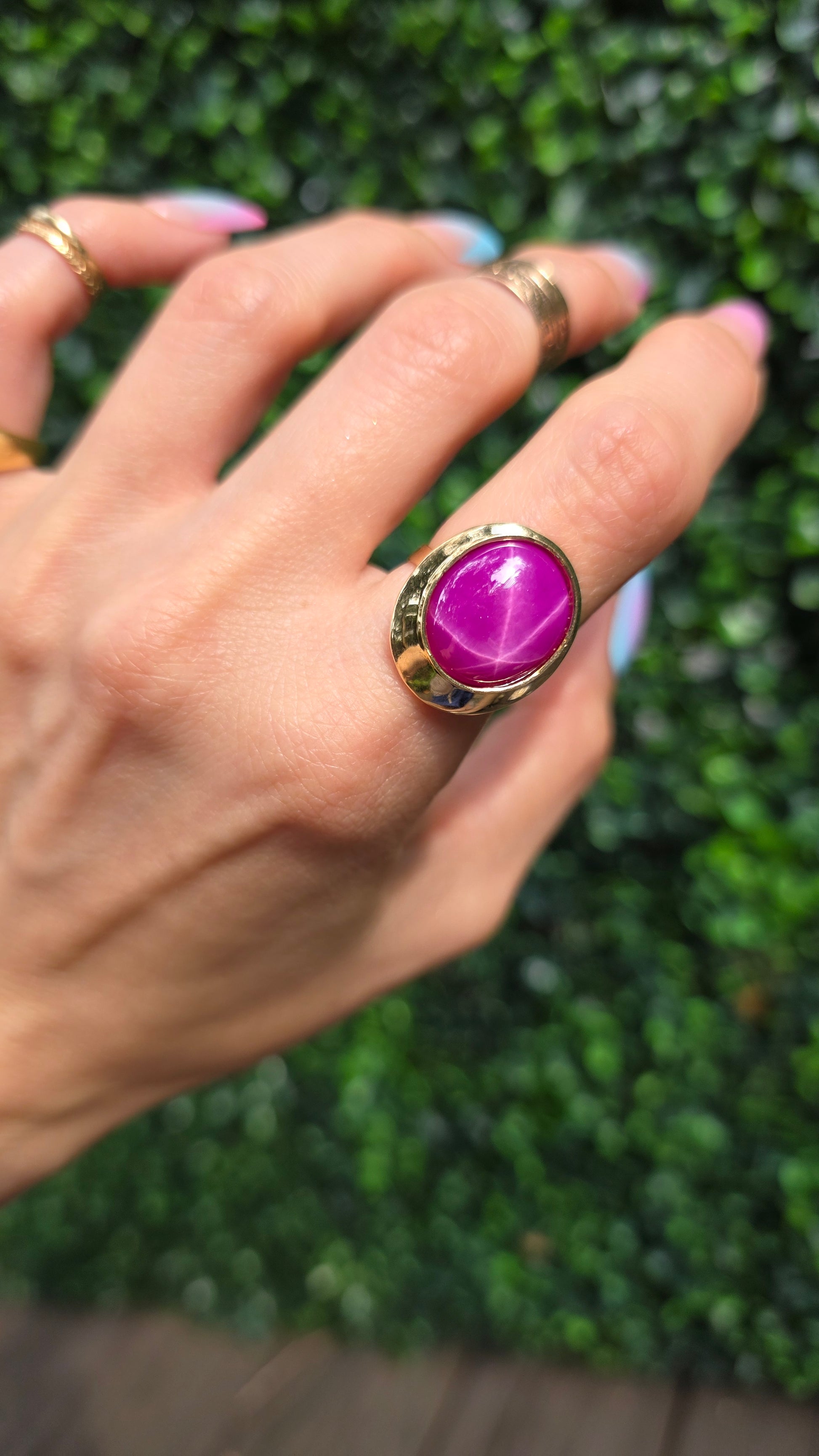 A gold ring with a large oval purple cabochon gemstone worn on a person's finger, with a green hedge in the background.