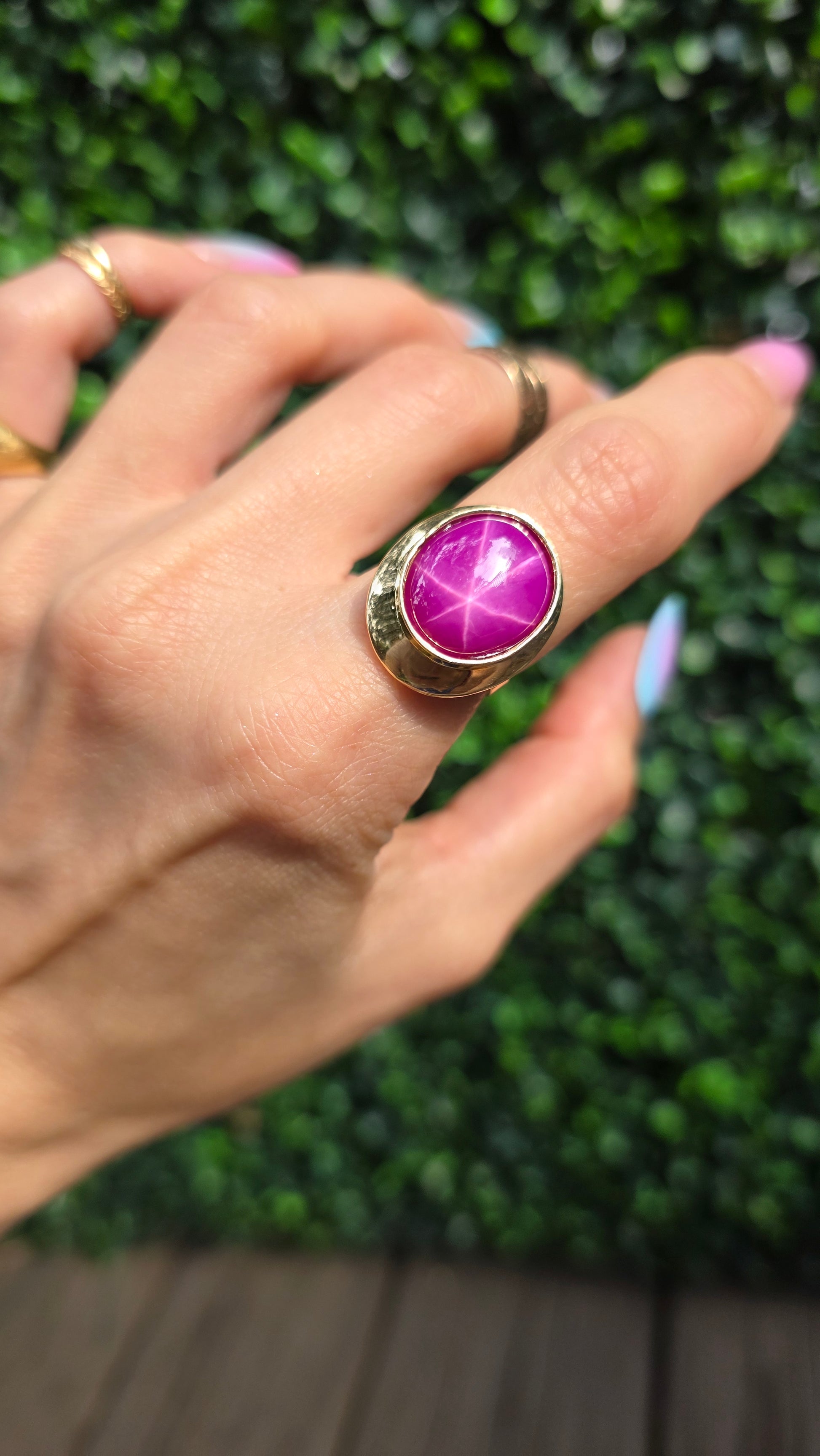 A gold ring with a large oval magenta-purple starburst cabochon gemstone, worn on a person's hand against a green hedge background.