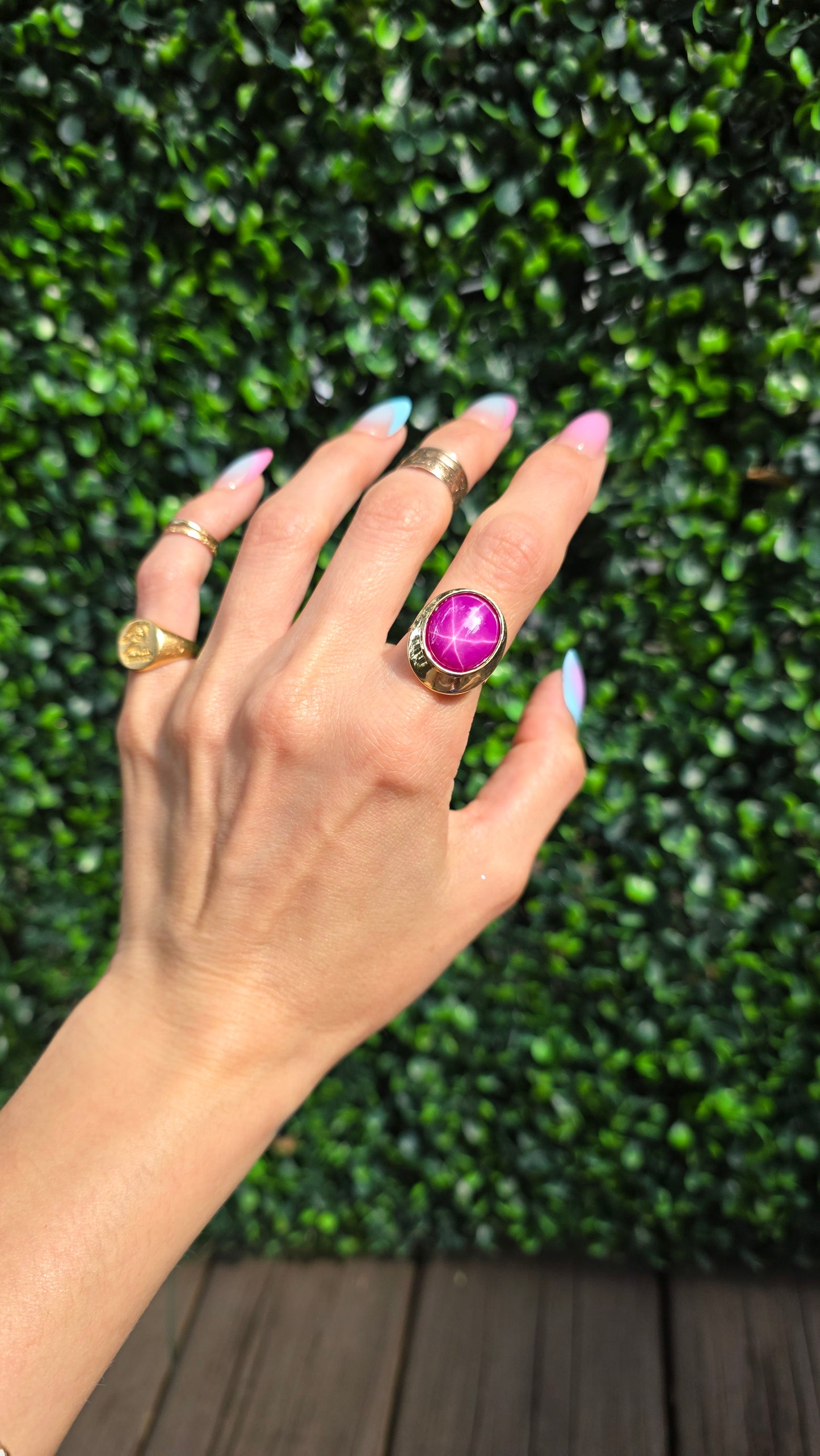 A gold ring with a large magenta-purple oval stone is worn on a person's hand, with other gold rings also visible, set against a green hedge background.