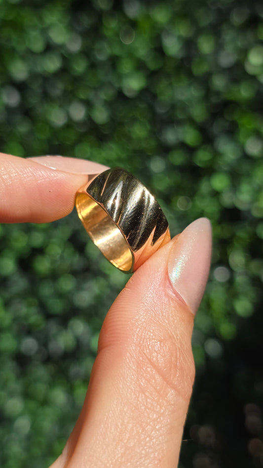 A close-up of a hand holding a wide, chunky gold ring with a textured, ribbed surface. The background is a soft-focus green hedge.