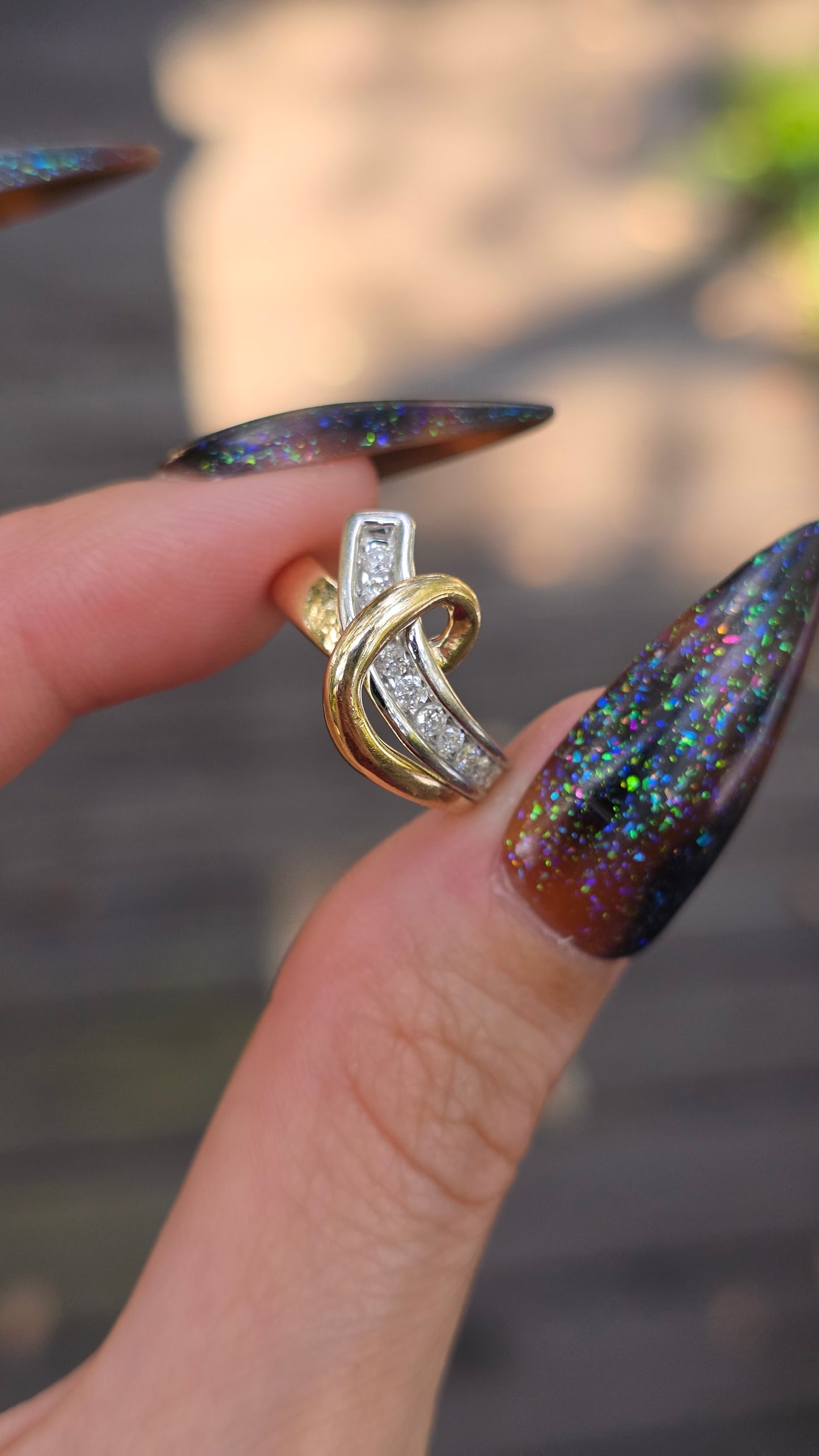 A close-up of a hand with long, dark glittery manicured nails holding a gold and silver interlocking ring with clear stones, set against a blurred outdoor background.