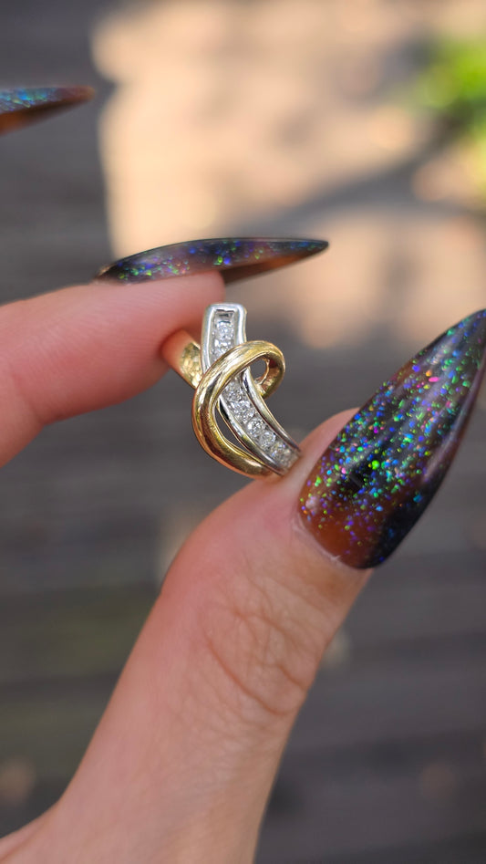 A close-up of a hand with long, dark glittery manicured nails holding a gold and silver interlocking ring with clear stones, set against a blurred outdoor background.